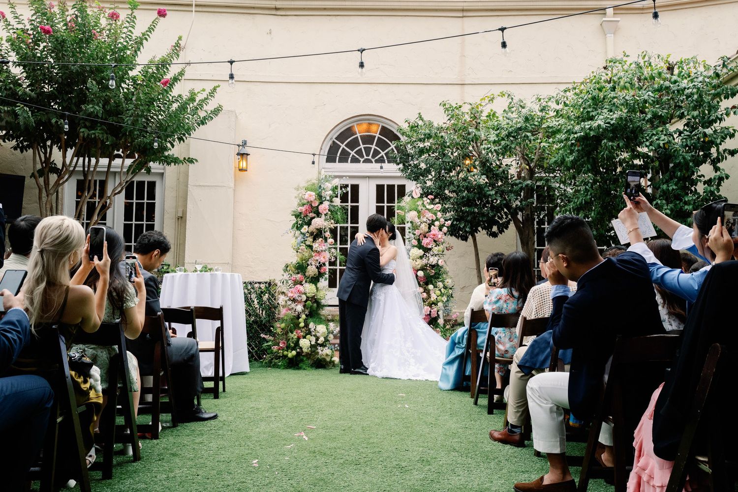 Bride and groom kiss at outdoor garden wedding ceremony with floral arch and seated guests.