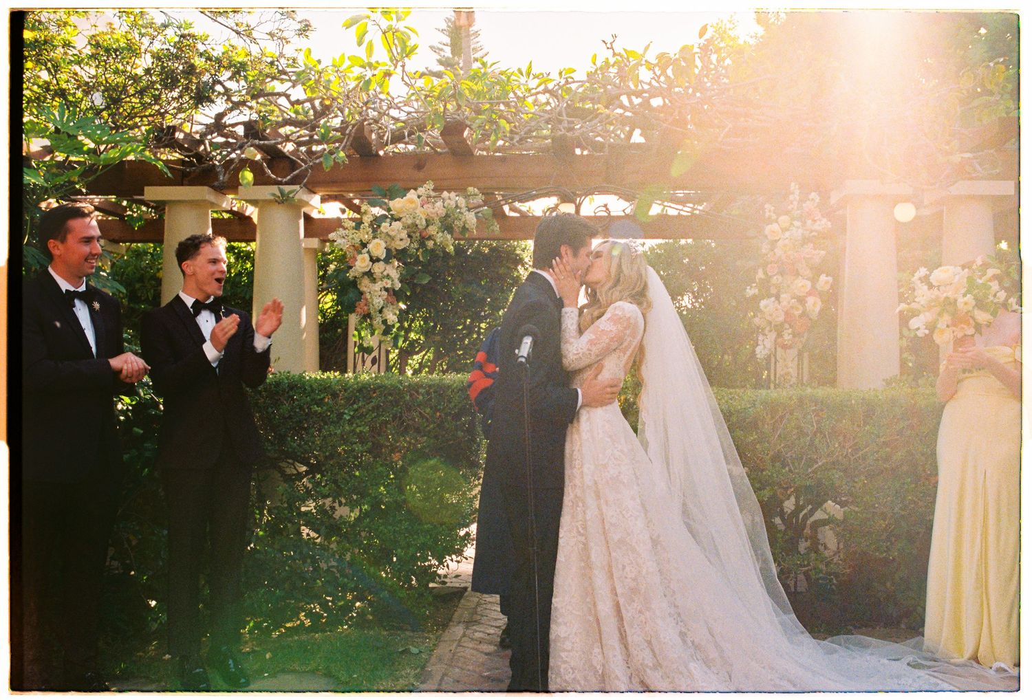 Bride and groom share first kiss under sunlit floral pergola as wedding guests cheer and applaud.