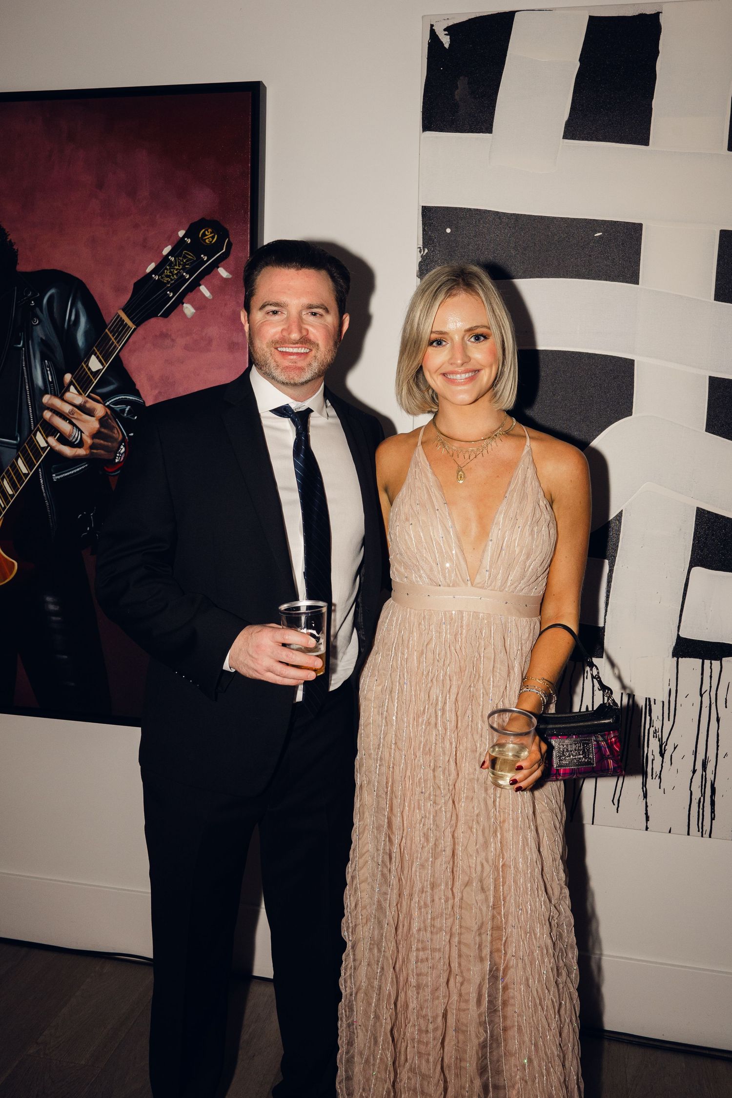 Elegant couple posing at an art gallery event, man in suit and woman in blush gown holding drinks.