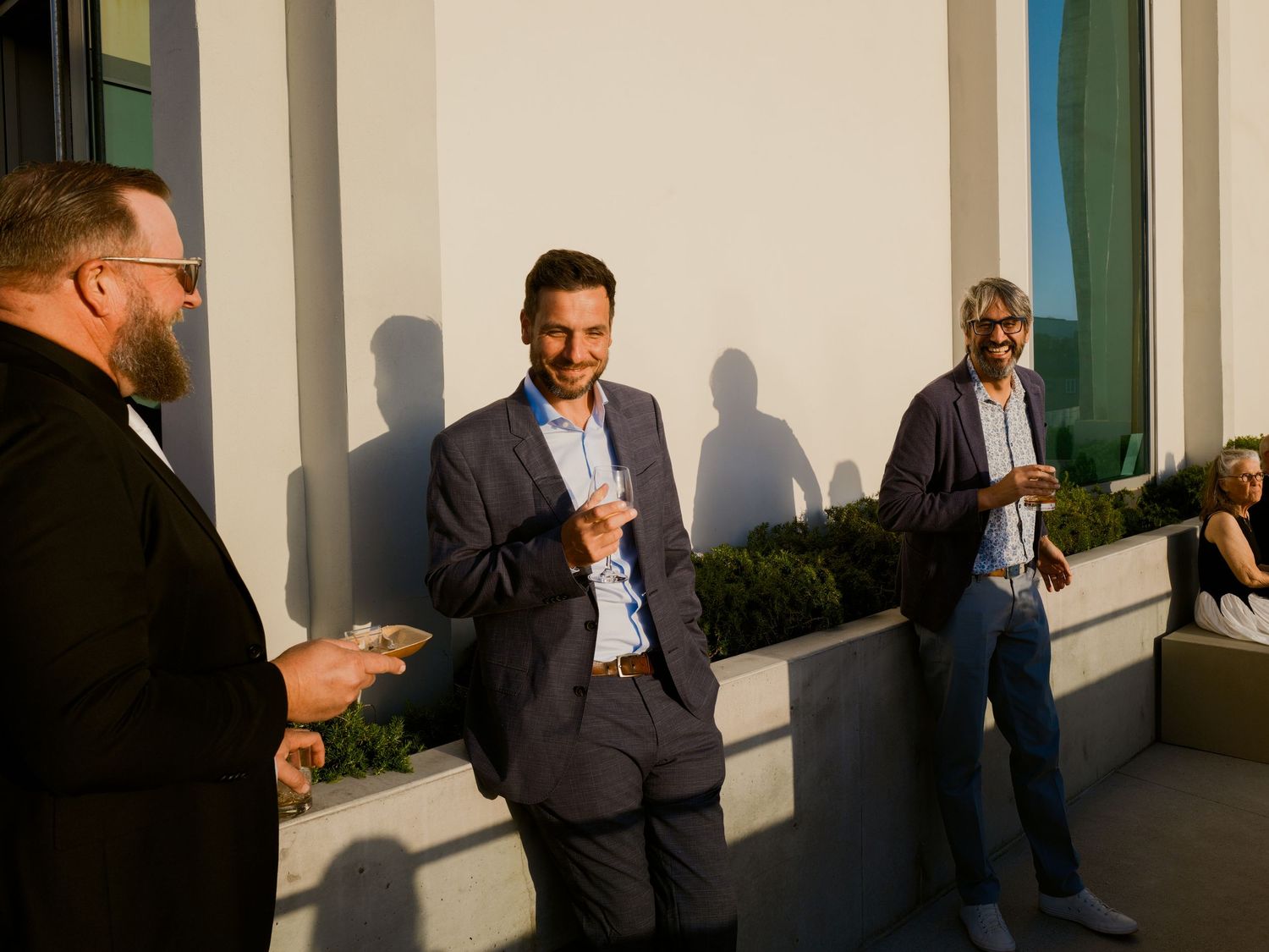 Three men in business casual attire laughing and socializing outdoors at a networking event in warm evening light.