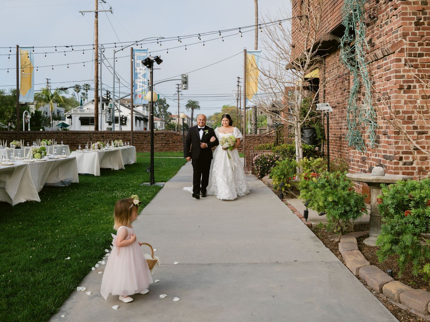 Bride and groom walking down outdoor aisle with flower girl at garden wedding venue.