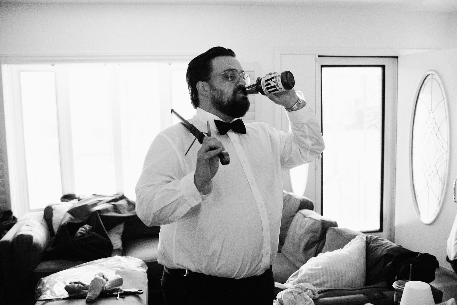 Groom in bow tie and suspenders drinks from can while holding razor during wedding preparations.
