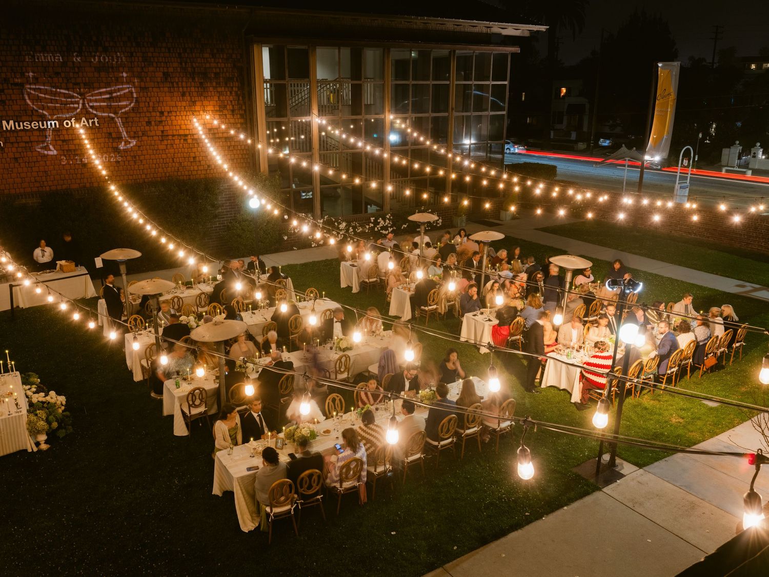Aerial view of elegant outdoor wedding reception at night with string lights and round tables on a lawn.