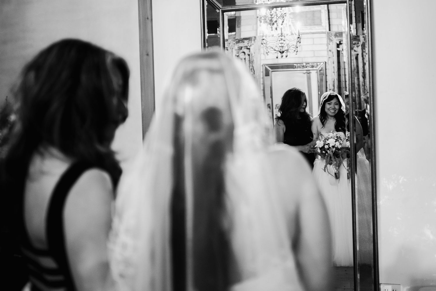 Bride in white gown and veil admires her reflection in an ornate mirror during wedding preparations.