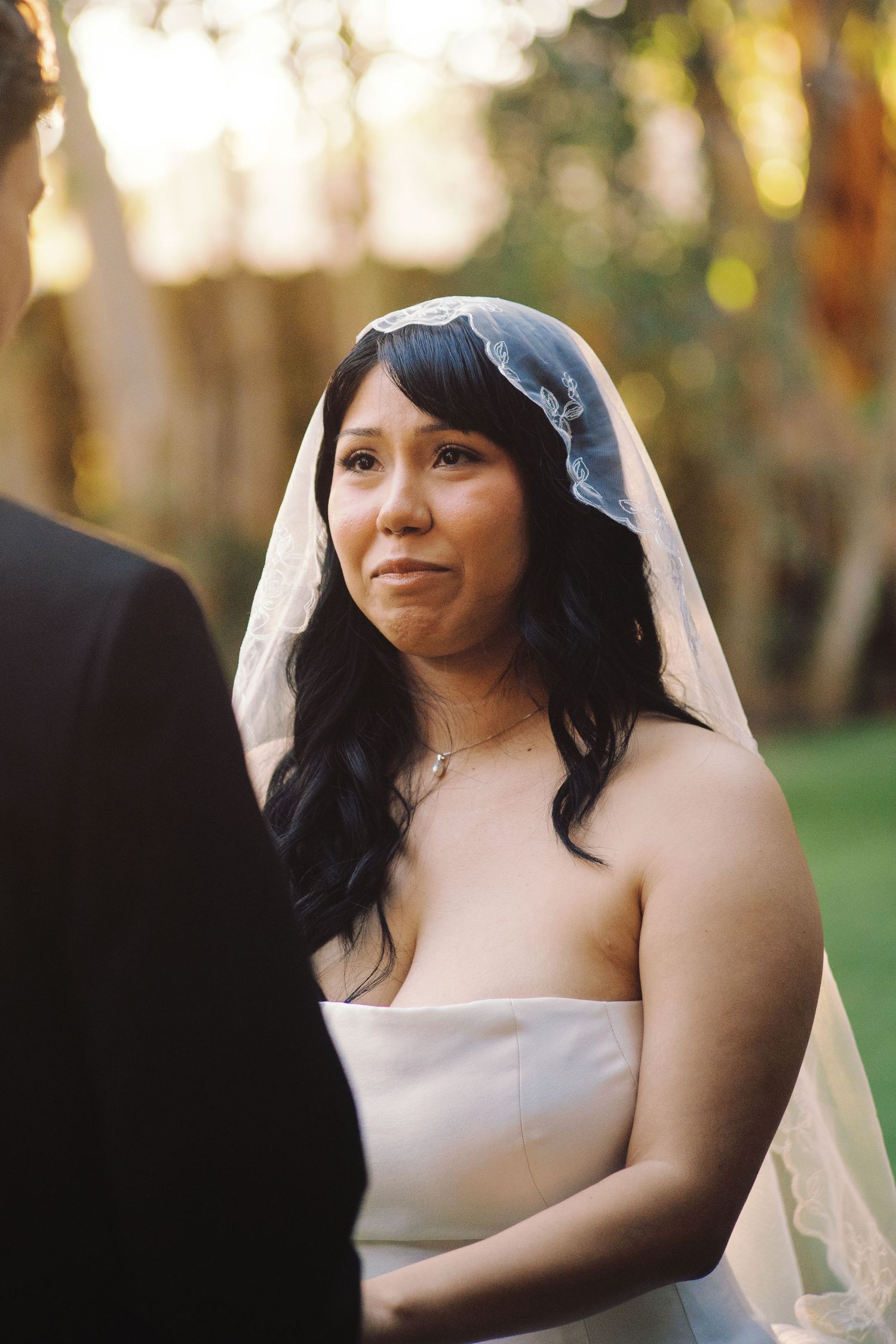 Emotional bride in white strapless gown and lace veil crying during outdoor wedding ceremony at golden hour.