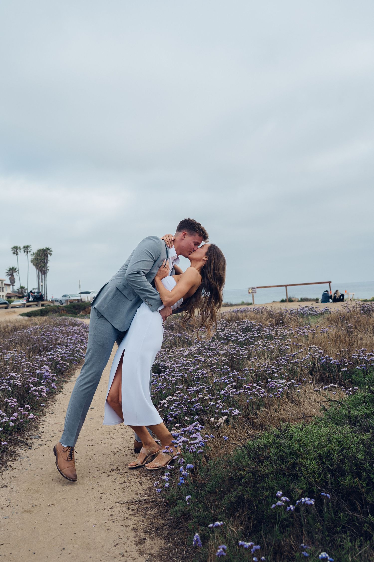 Couple sharing a romantic dip kiss on a wildflower-lined coastal path, man in gray suit and woman in white strapless dress.