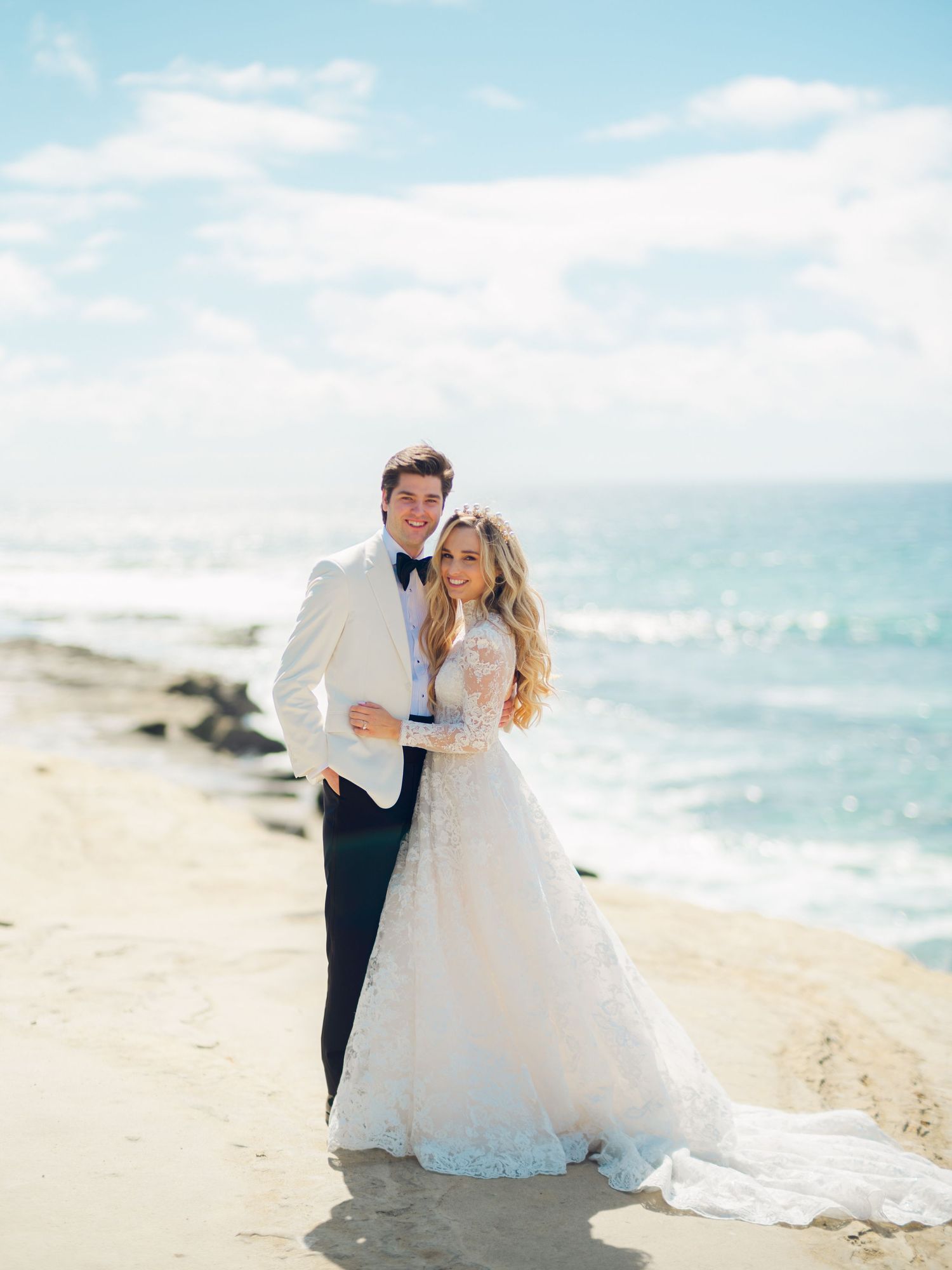 Bride and groom embrace on a sandy beach, with ocean waves and blue sky behind them.