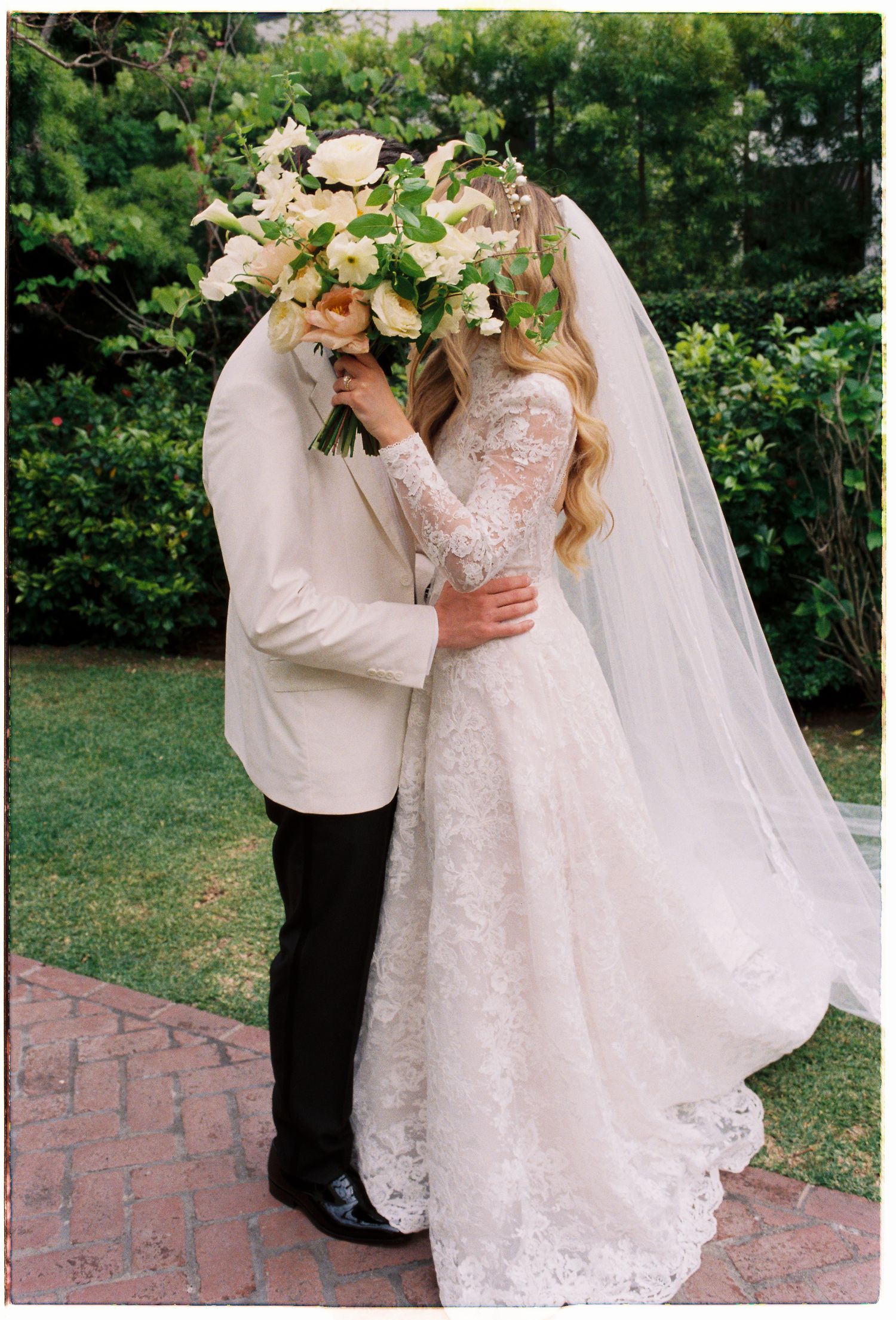 Bride and groom share a romantic kiss hidden behind a lush floral bouquet in a garden setting.