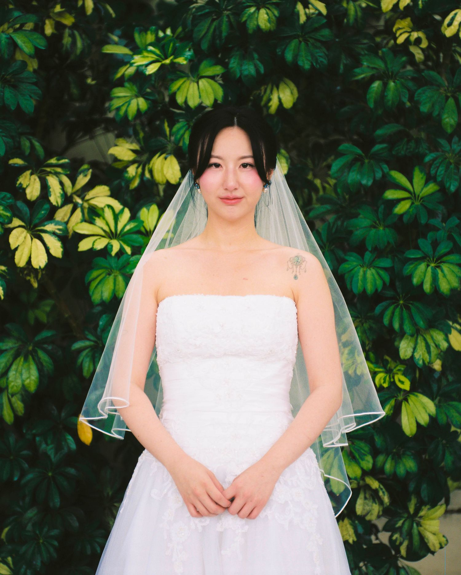 Bride in strapless white lace gown and veil posing before lush greenery.