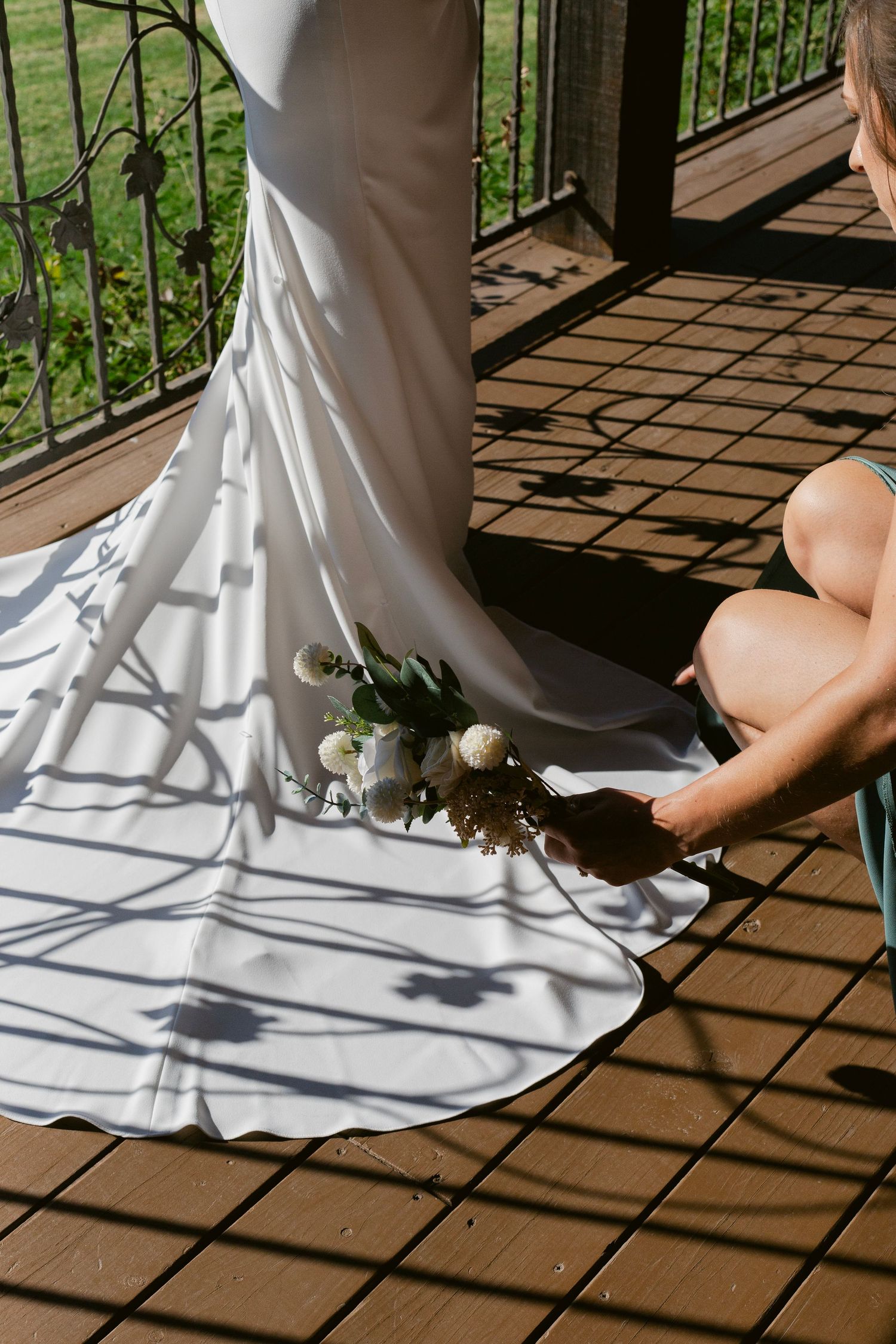 Bridesmaid arranging the train of a white wedding dress on a sunlit wooden deck with decorative iron fence shadows.