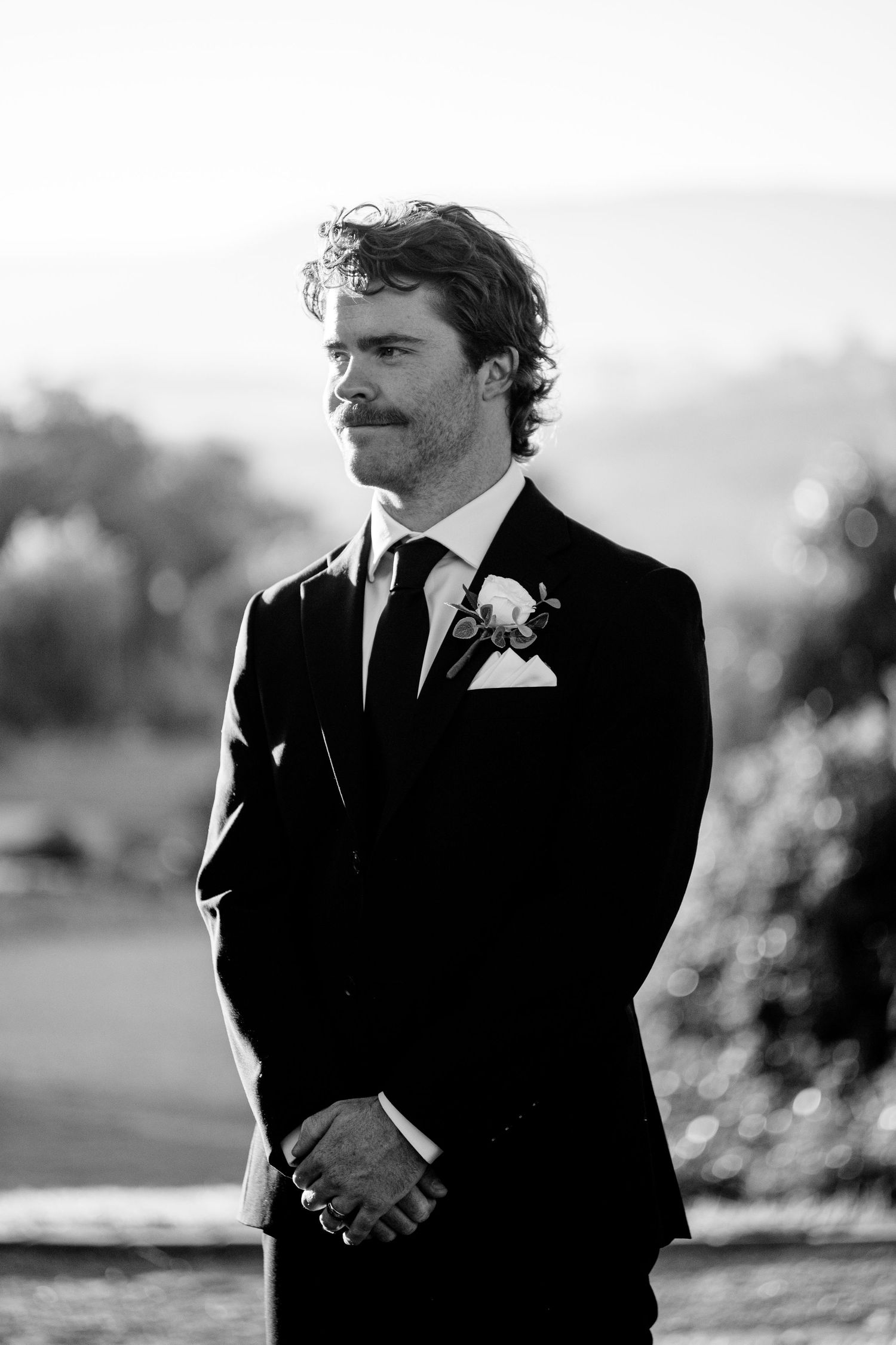 Dapper groom in black tuxedo with boutonniere stands confidently in a classic black and white portrait.