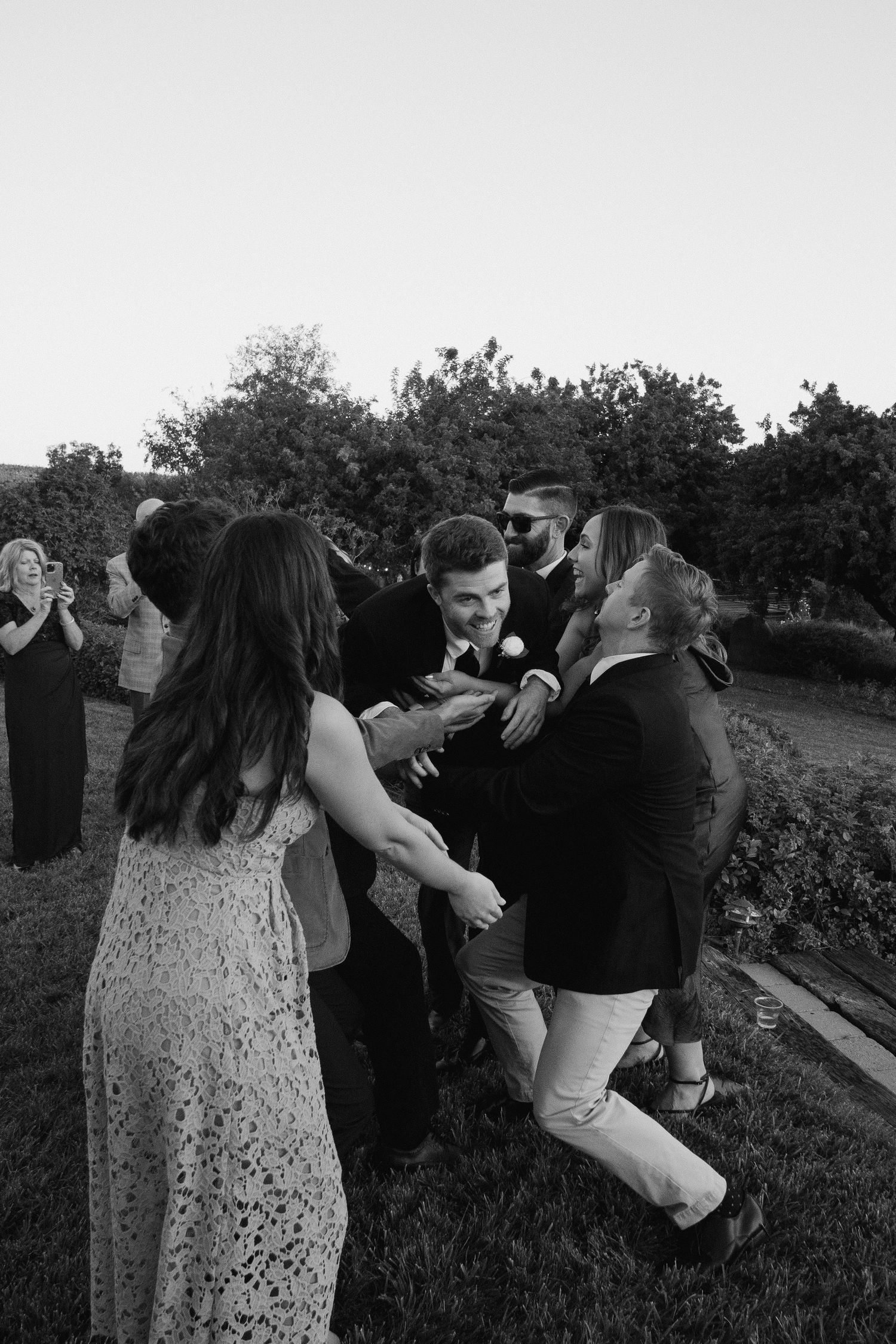 Black and white photo of a groom laughing as friends playfully surround him during outdoor wedding cocktail hour.
