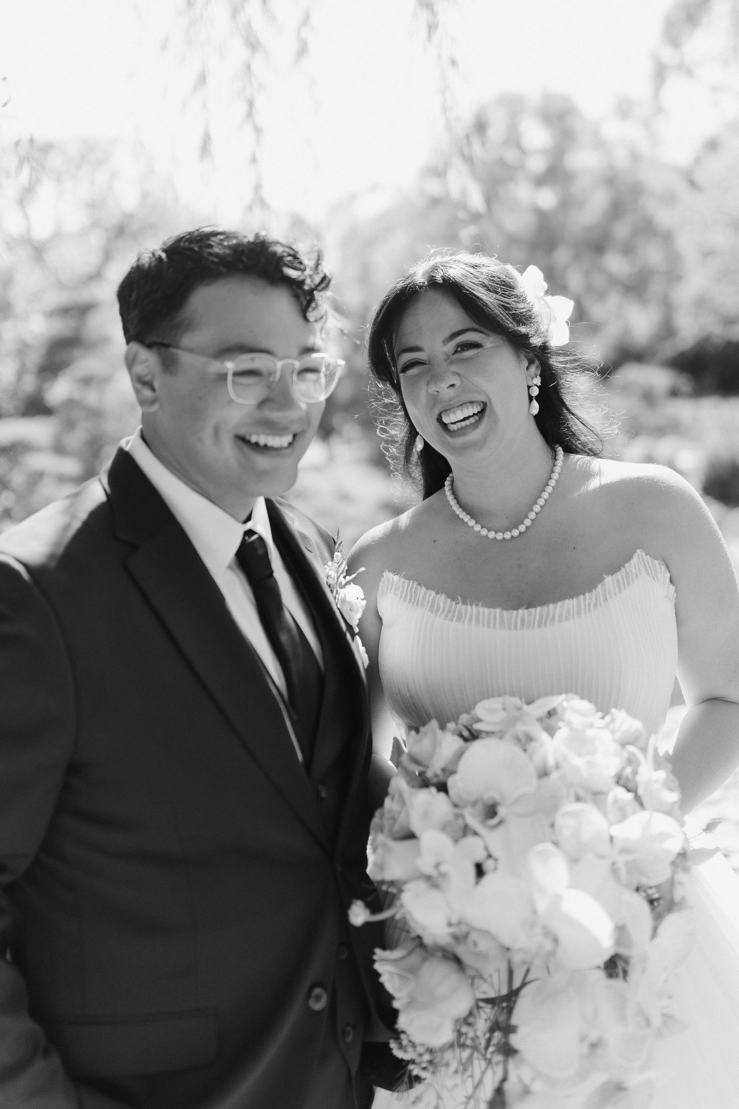 Black and white photo of smiling bride holding bouquet and groom in suit at outdoor wedding ceremony.