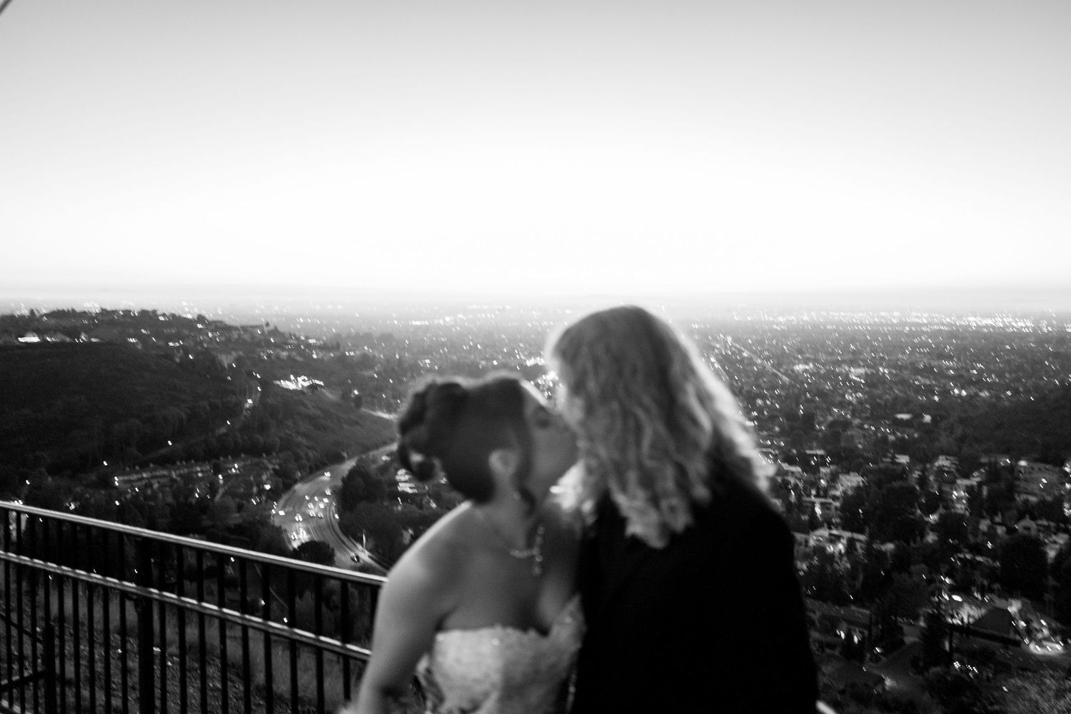 Black and white photo of a couple sharing a kiss at their wedding with a city skyline in the background.