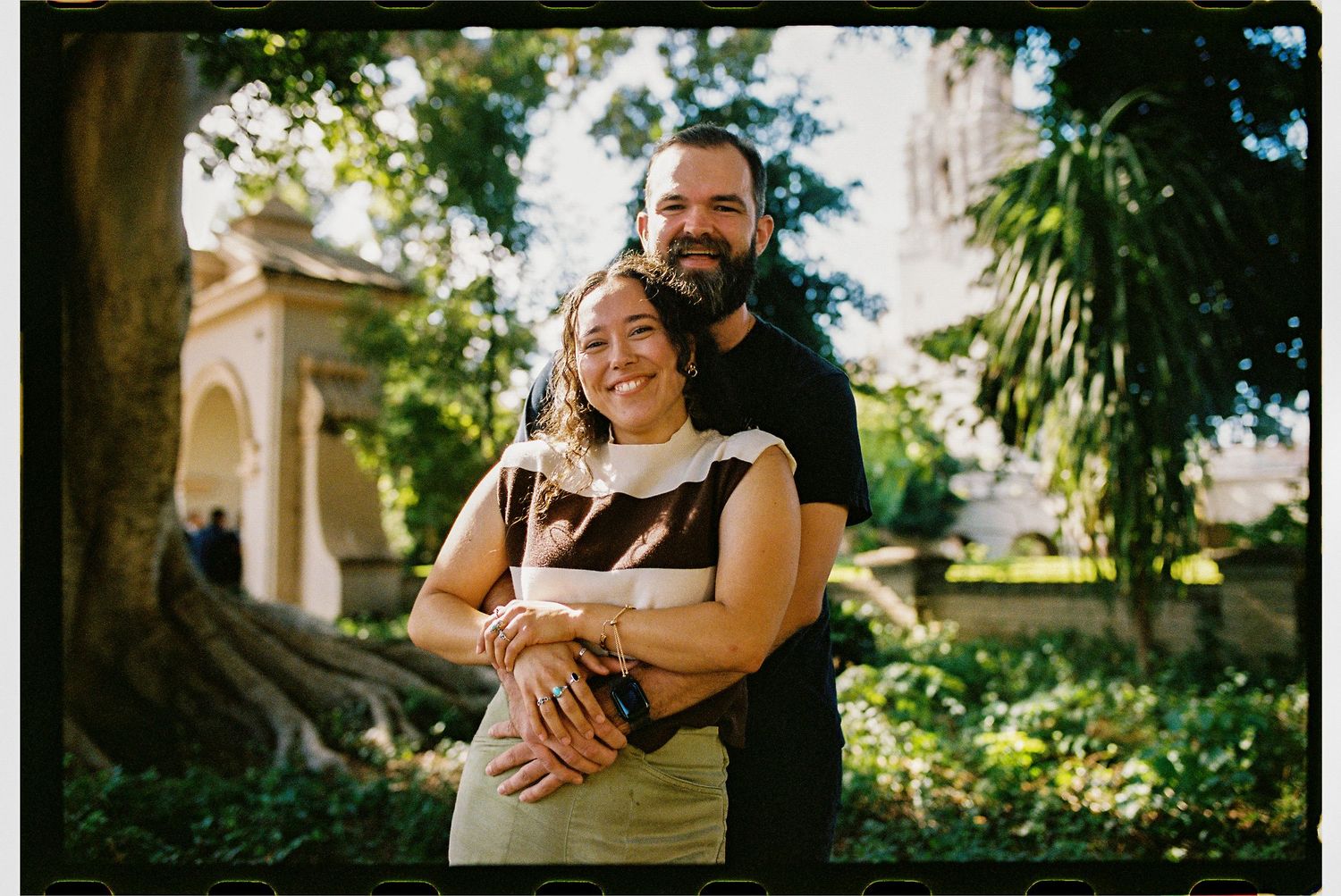 A smiling couple embraces in a lush garden with historic architecture and trees in the background.