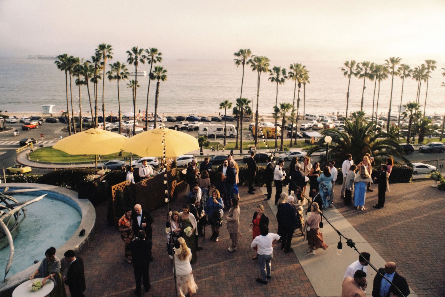 Aerial view of wedding cocktail hour on oceanfront terrace with palm trees and harbor views at sunset.