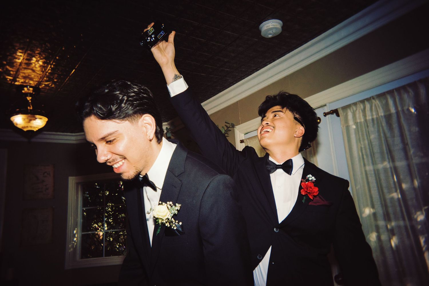 Two groomsmen in black tuxedos celebrate joyfully, one raising a trophy in a warmly lit room.