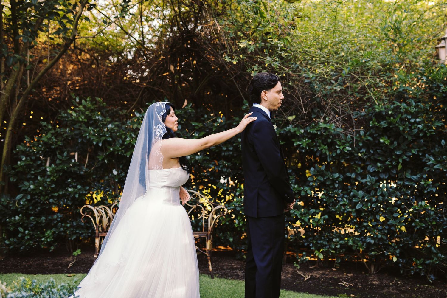 Bride in white gown and veil tapping groom's shoulder during first look outdoors.