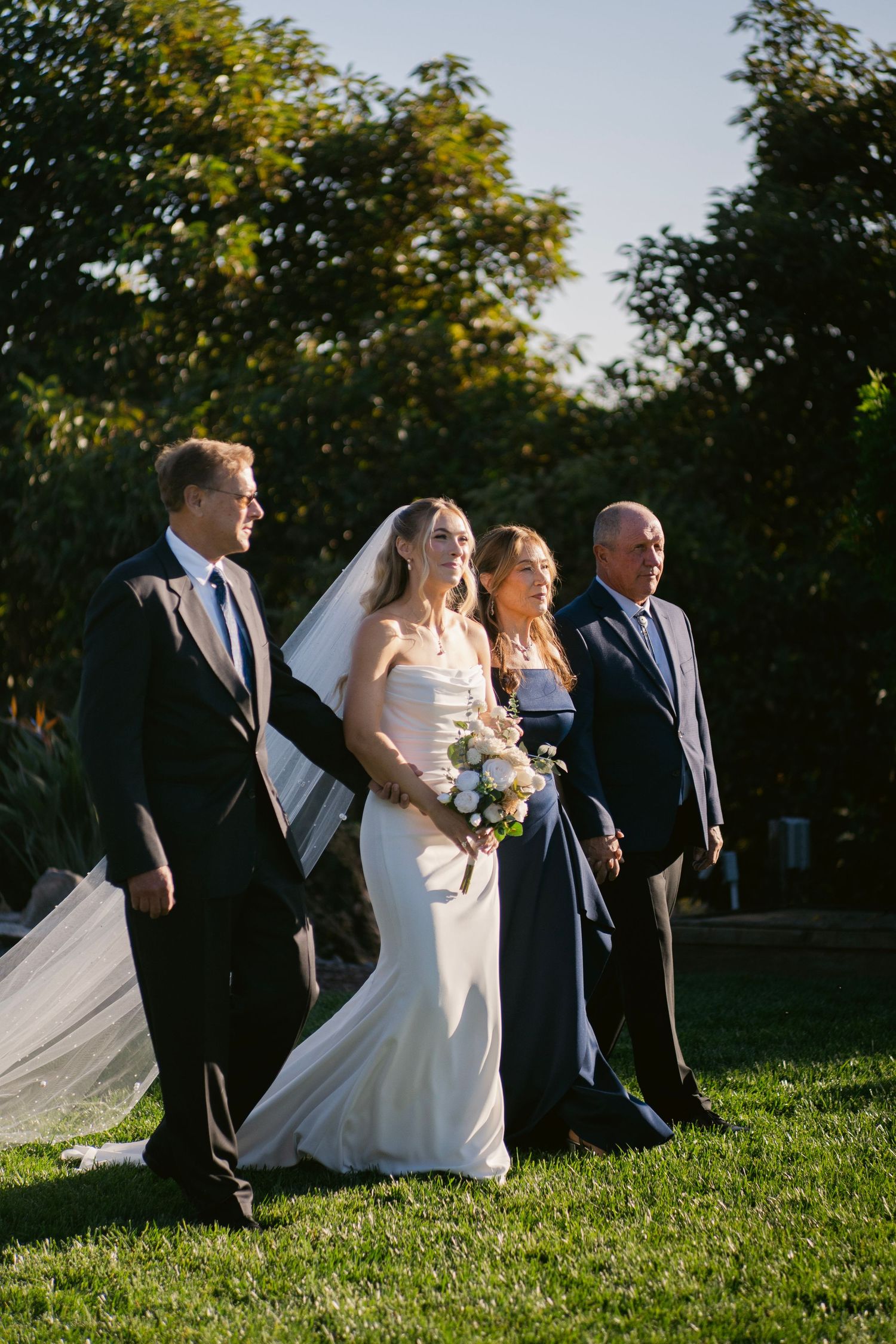 Bride with bouquet walks down aisle escorted by parents in elegant outdoor wedding ceremony.