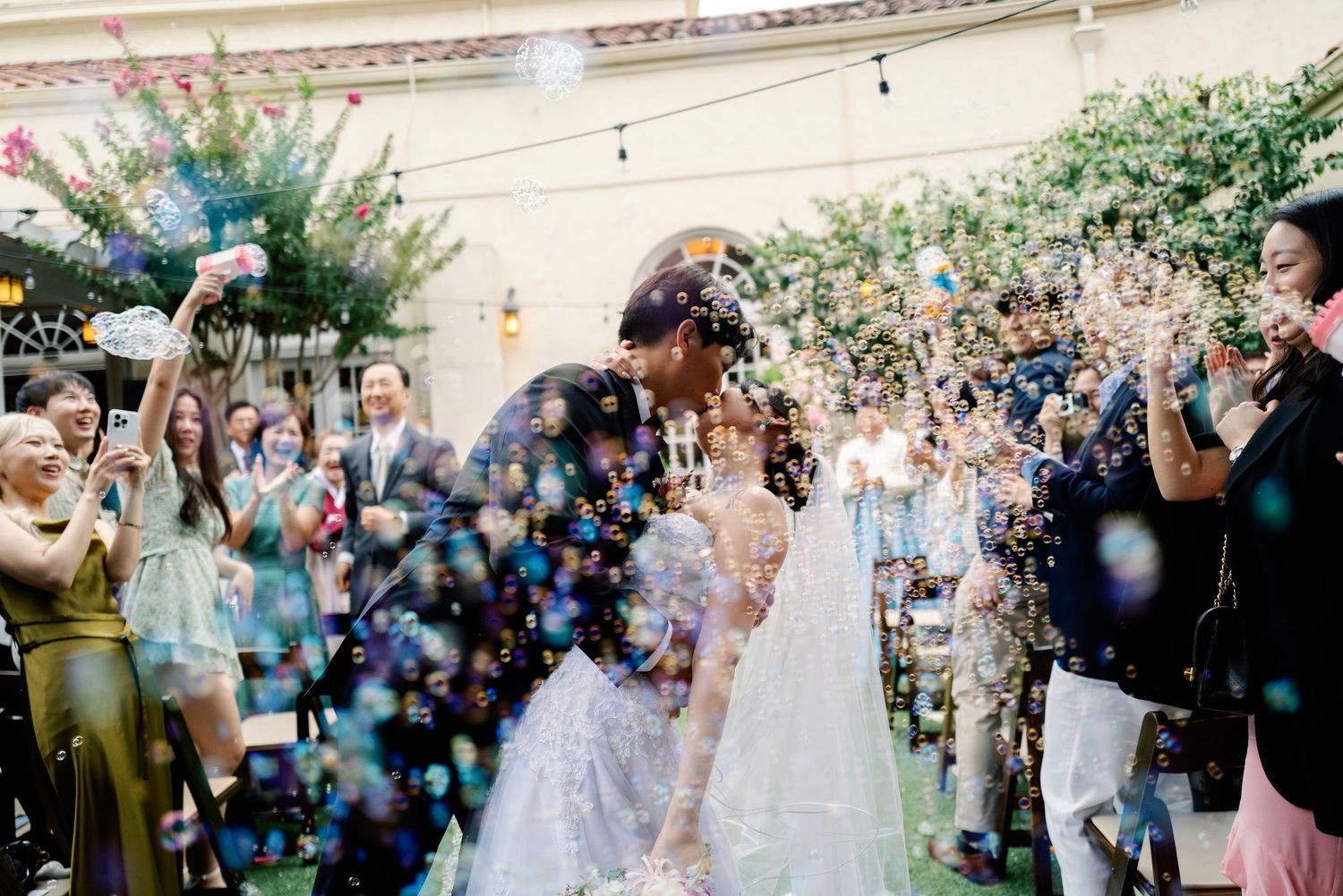 Bride and groom celebrate recessional with joyful bubble shower and cheering guests at garden wedding.