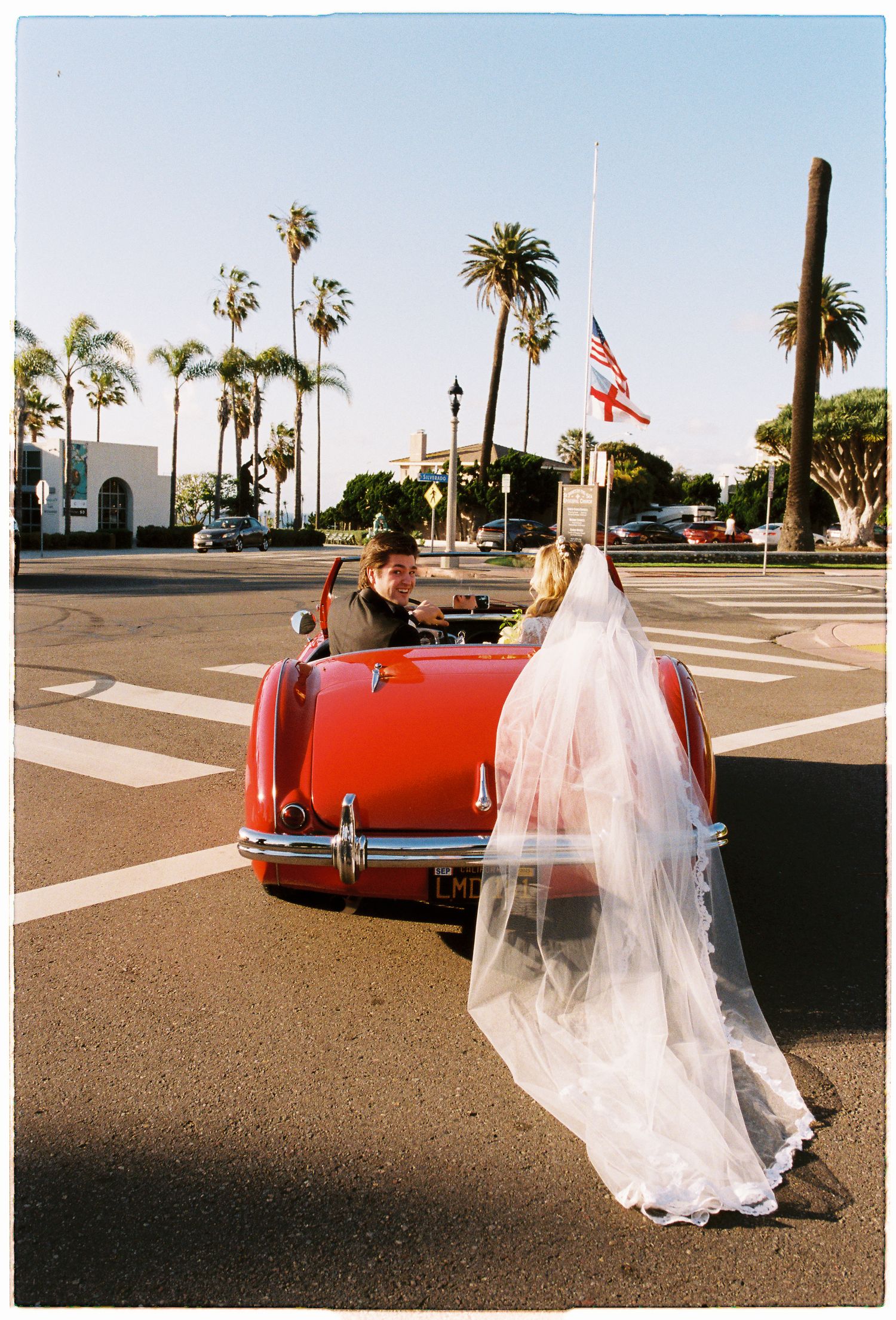 Bride in white gown and veil stands beside a classic red convertible car on a sunny street lined with palm trees.