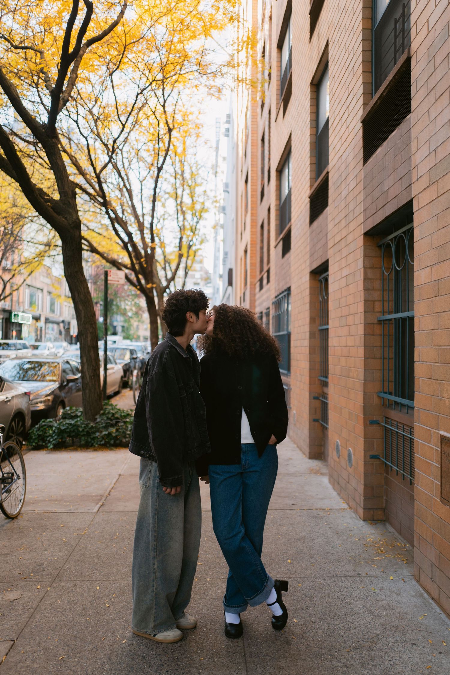 A couple shares a romantic kiss on an autumn city sidewalk surrounded by yellow trees and brick buildings.
