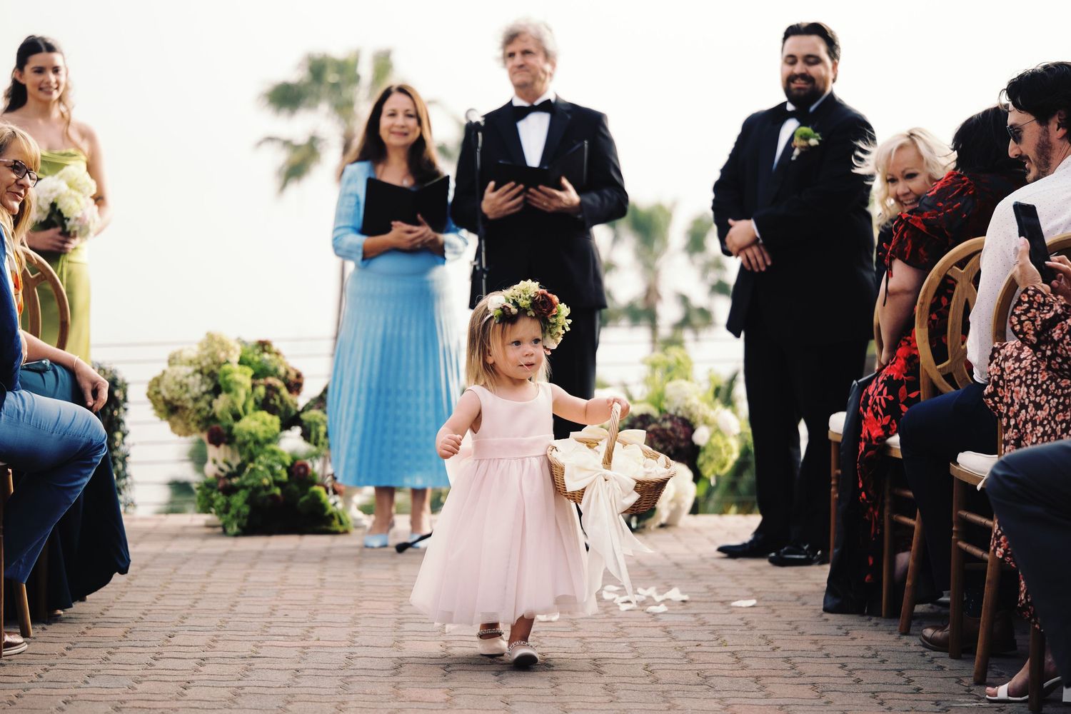 Adorable flower girl in pink dress walks down aisle at outdoor wedding ceremony.