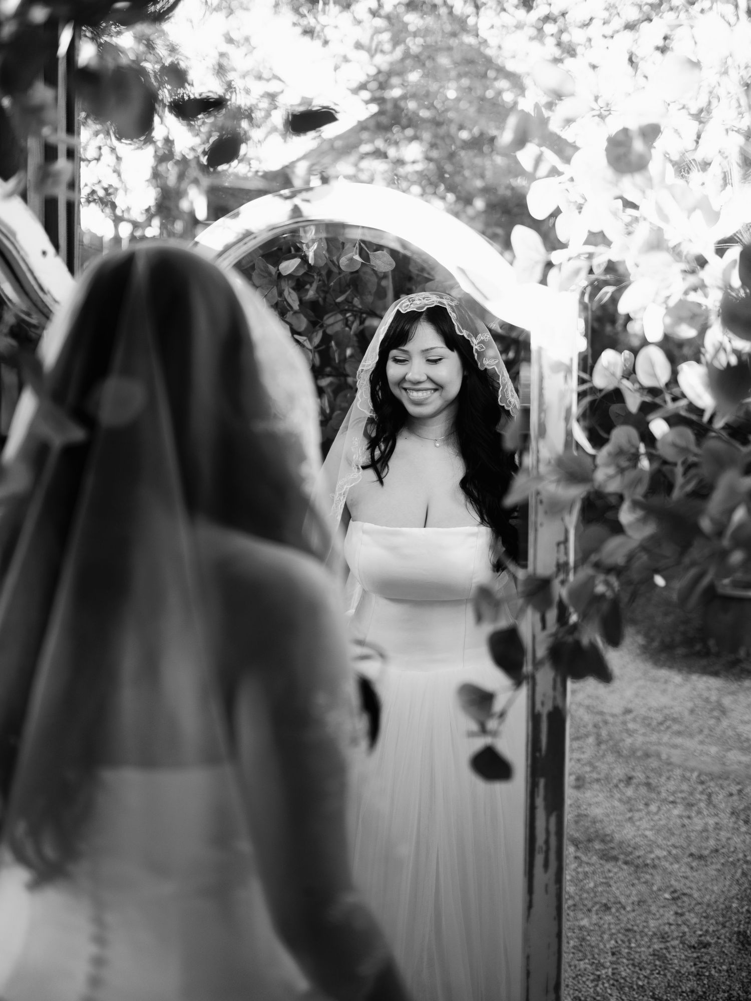 Smiling bride in white gown and veil views her reflection in an outdoor mirror surrounded by greenery.