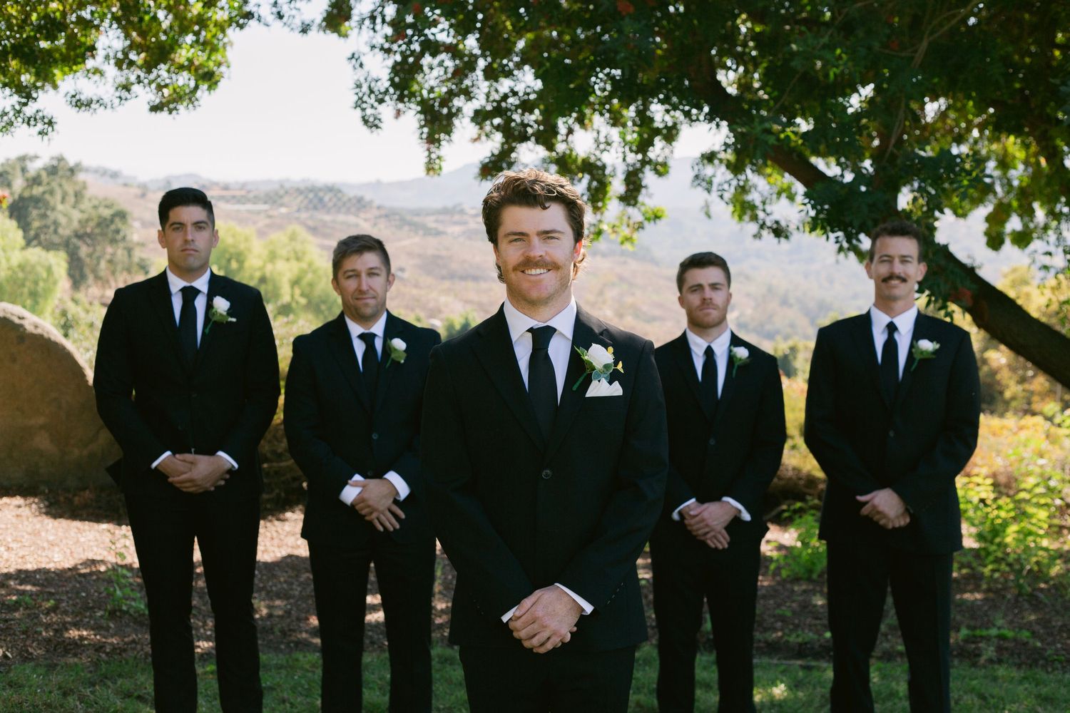 Groom smiling in a black suit with boutonniere, surrounded by four groomsmen under a tree with mountain backdrop.