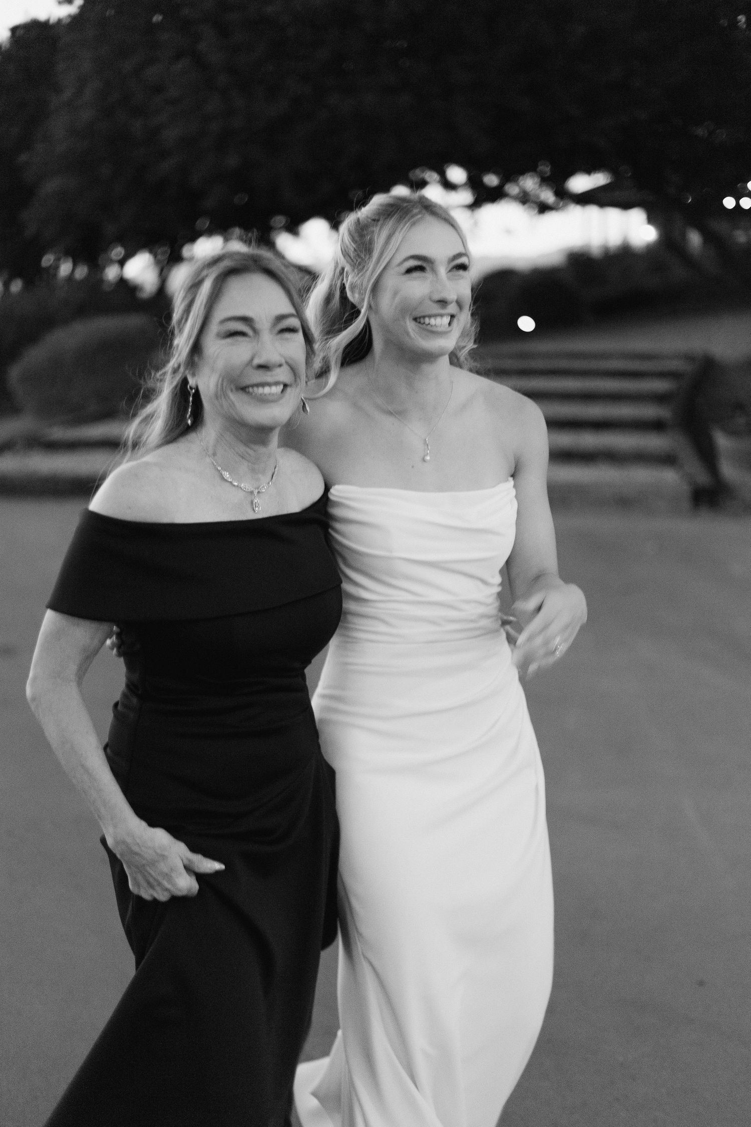 Black and white photo of smiling bride in white gown walking with mother in dark dress at wedding.
