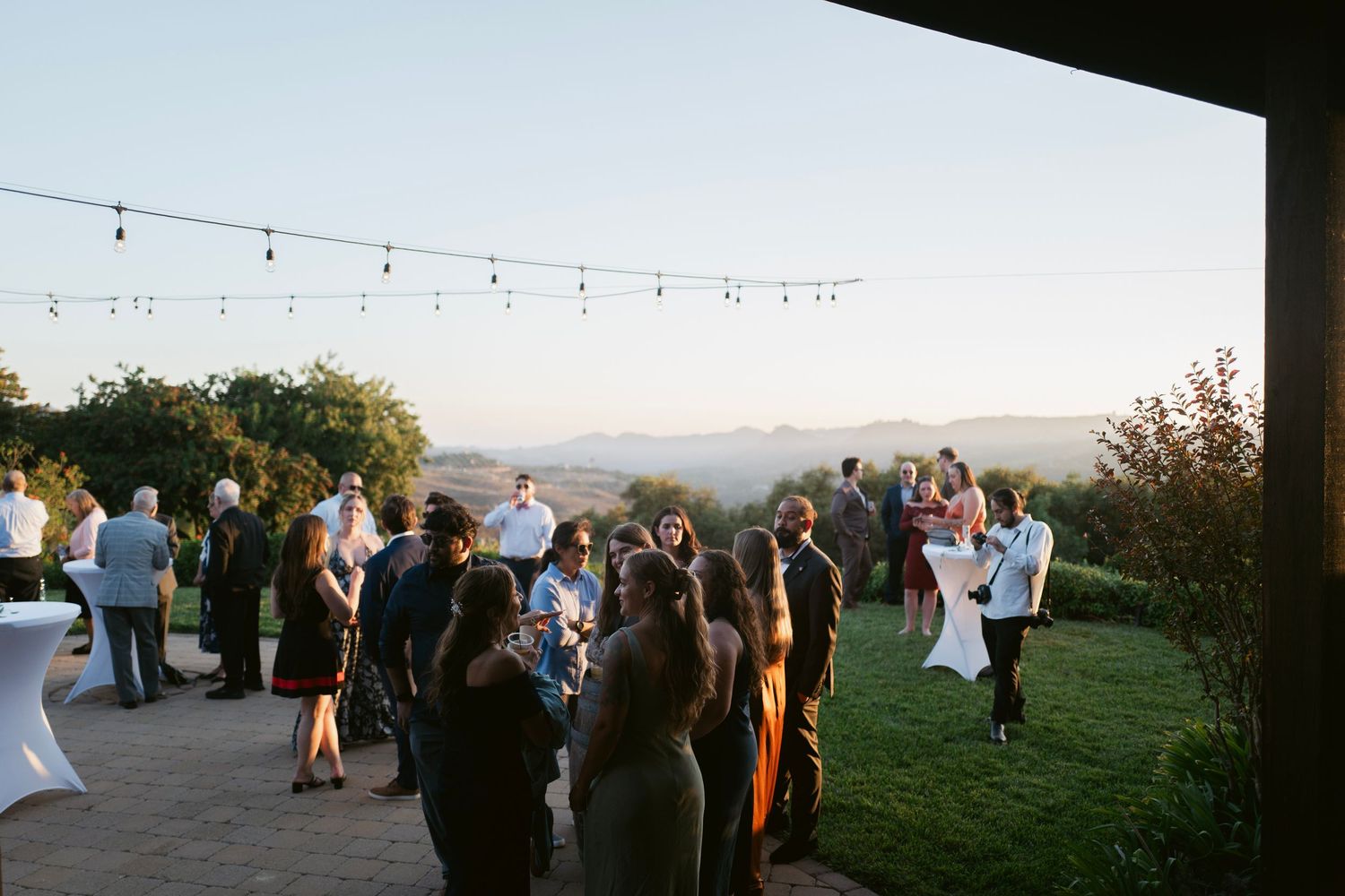Wedding guests mingle on a scenic outdoor patio with string lights and breathtaking mountain views at dusk.