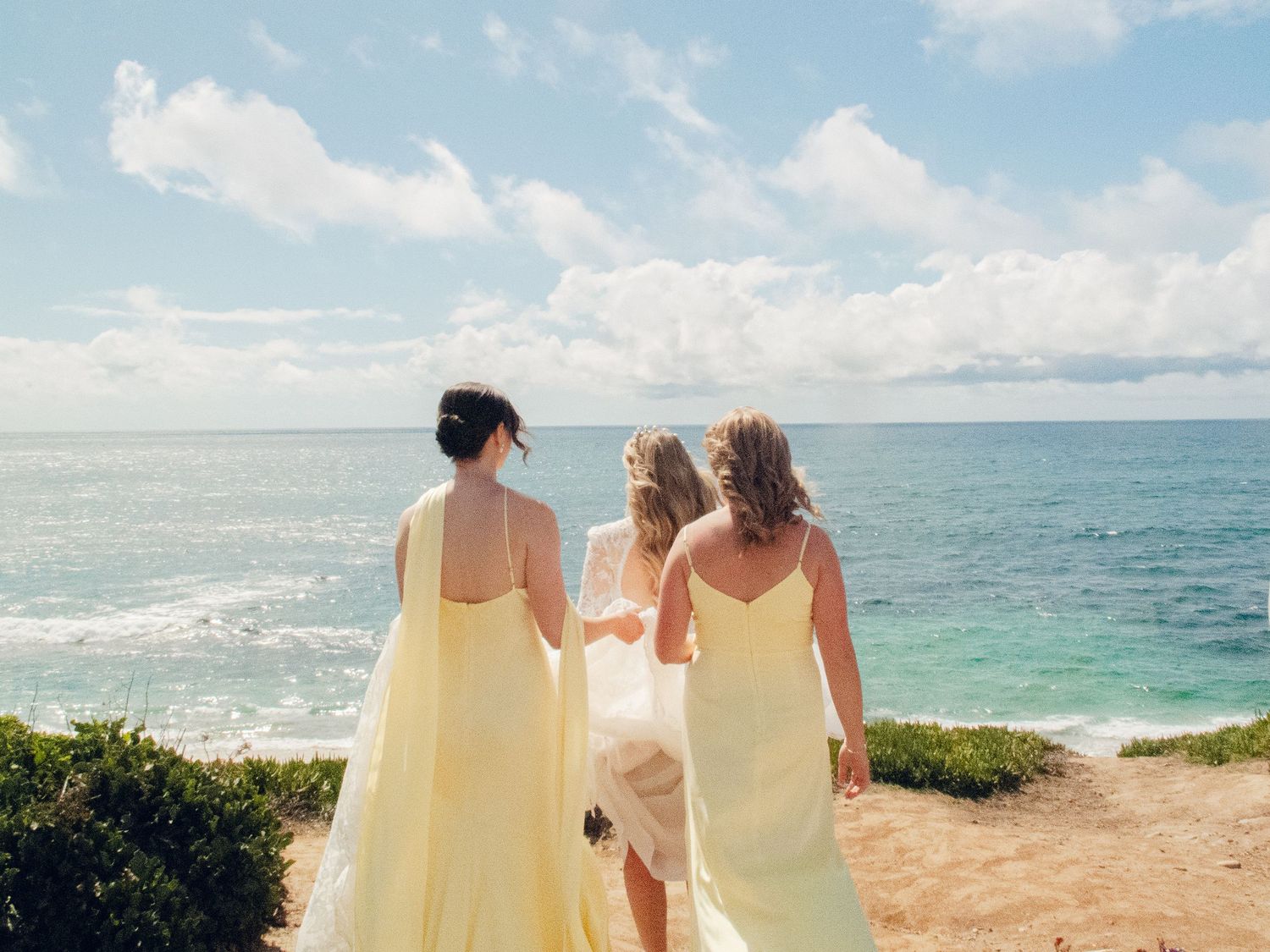 Three bridesmaids in yellow dresses walk toward turquoise ocean at a beach wedding.
