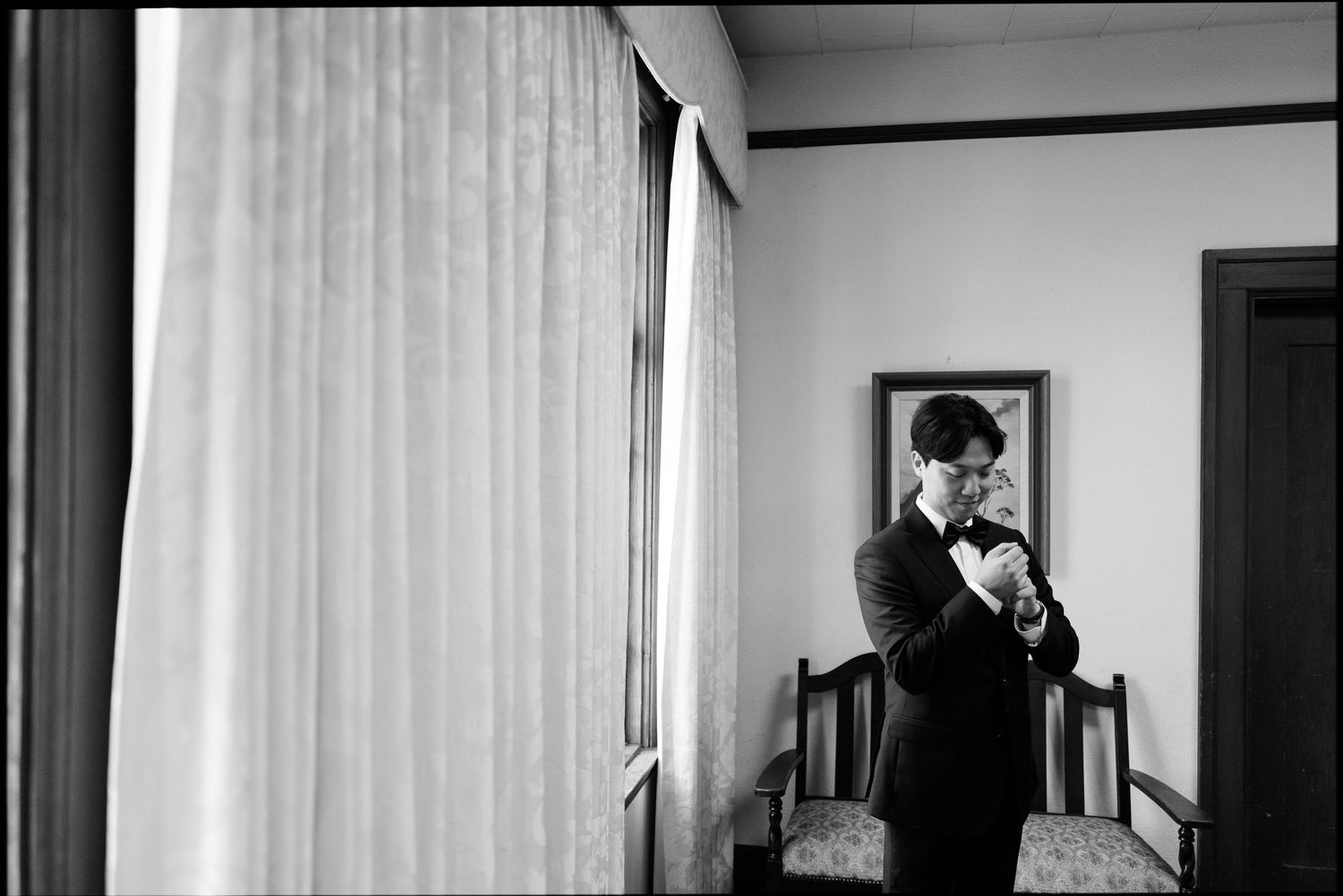 Black and white photo of groom adjusting bow tie by window before wedding.