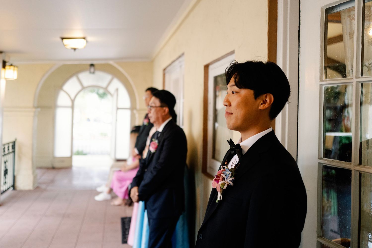 Groom in a tuxedo with boutonniere standing in a sunlit corridor before his wedding ceremony.