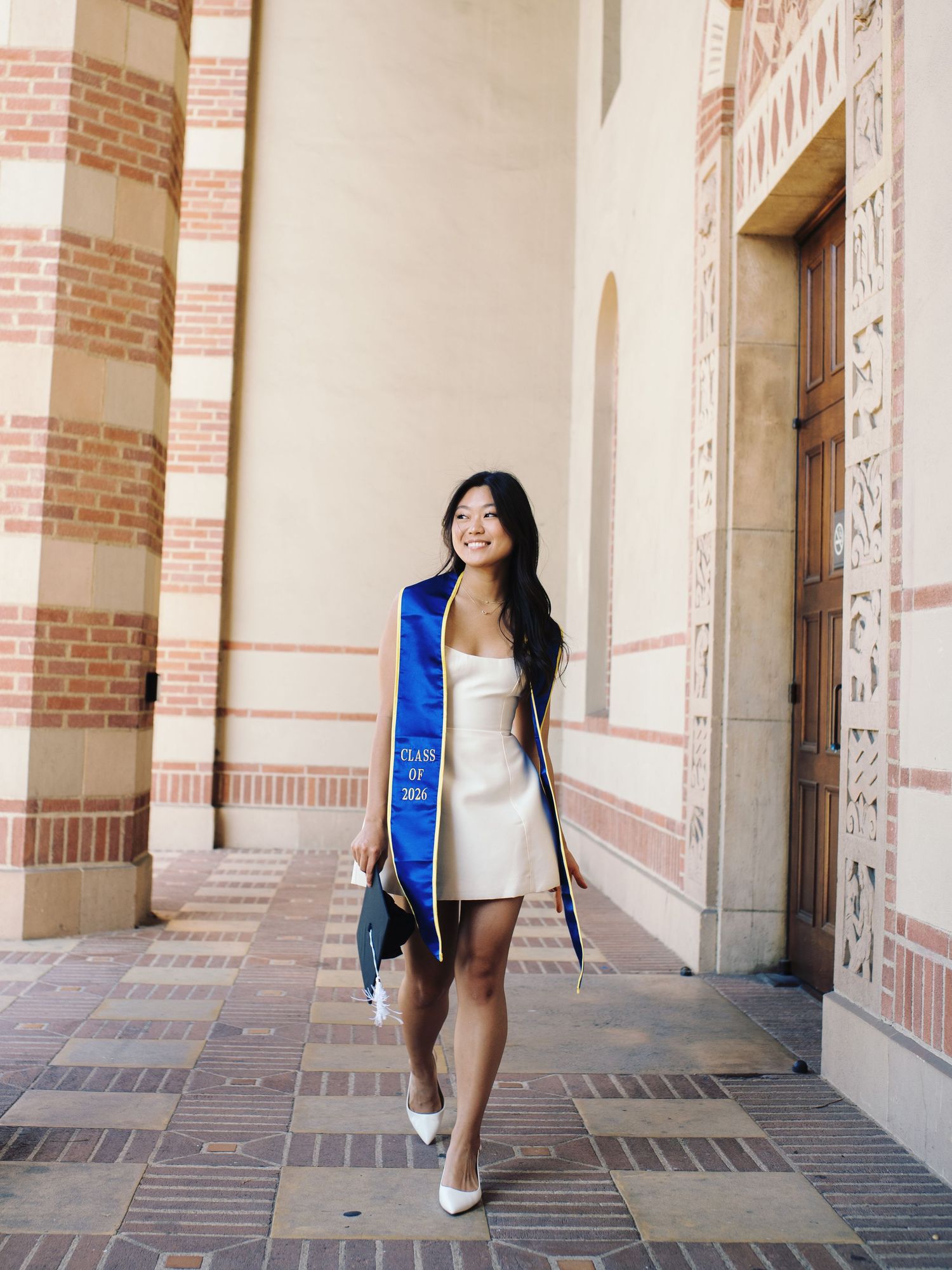 Graduate in white mini dress with blue UCLA stole walks through brick archway holding cap.