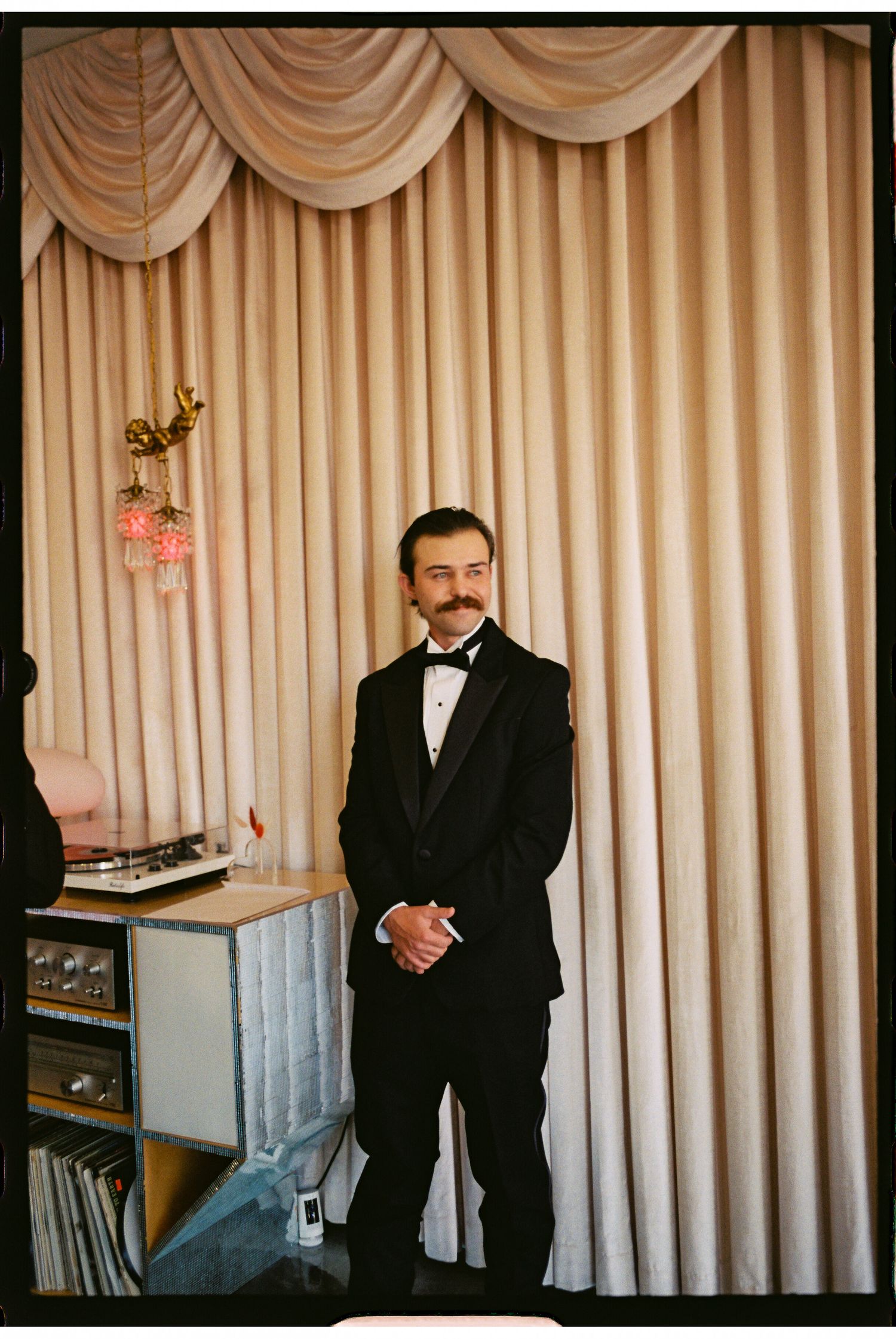 Man in black tuxedo with mustache standing before elegant cream draped curtains at formal event.