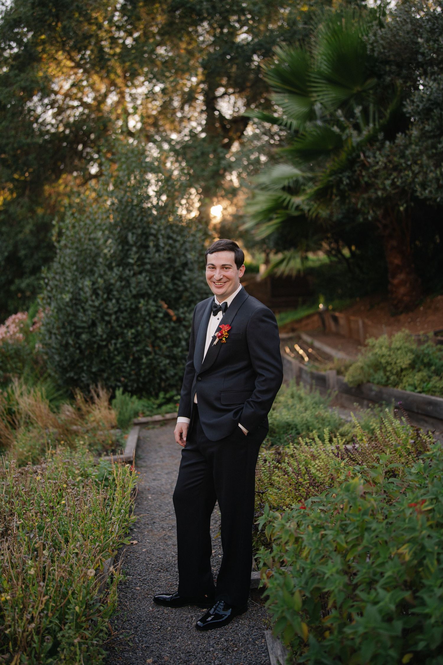Smiling groom in a black tuxedo with bow tie and red boutonniere stands on a gravel garden path surrounded by lush greenery at sunset.