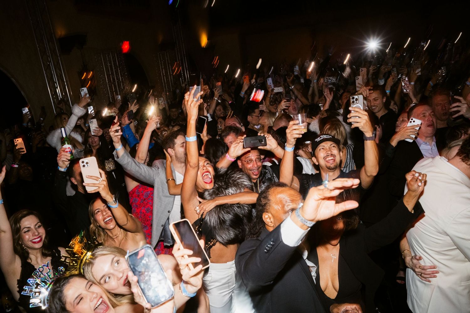 Crowd of excited party guests dancing and holding up phones with lights at a lively evening celebration.
