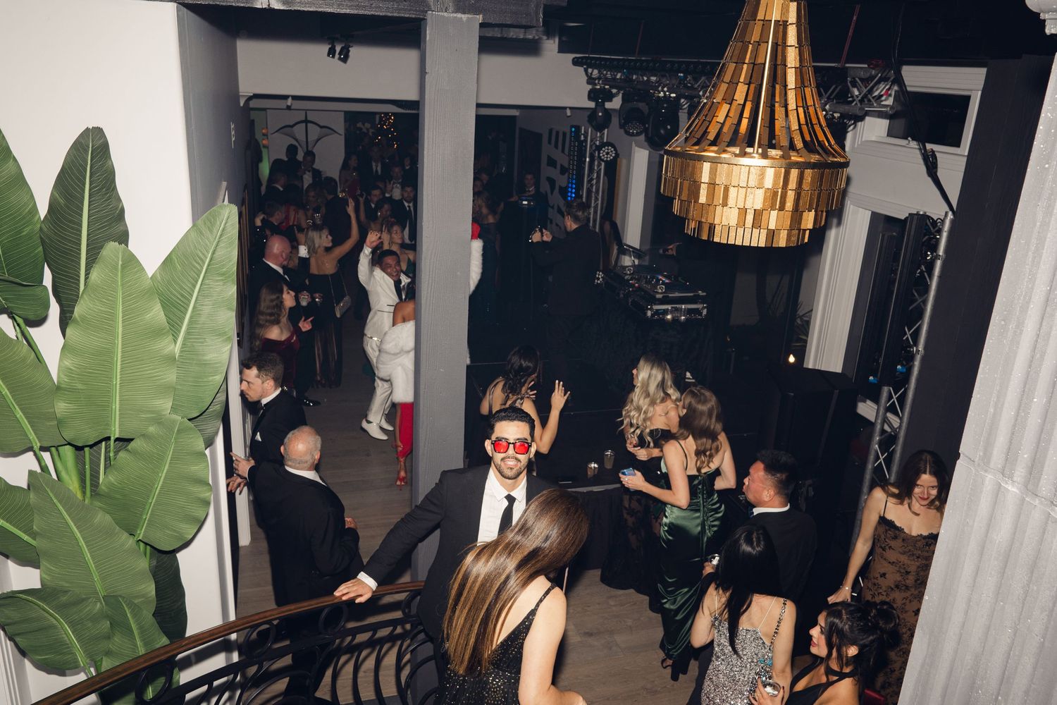 Elegant party guests in formal attire mingle inside a stylish venue with gold chandelier and tropical decor.