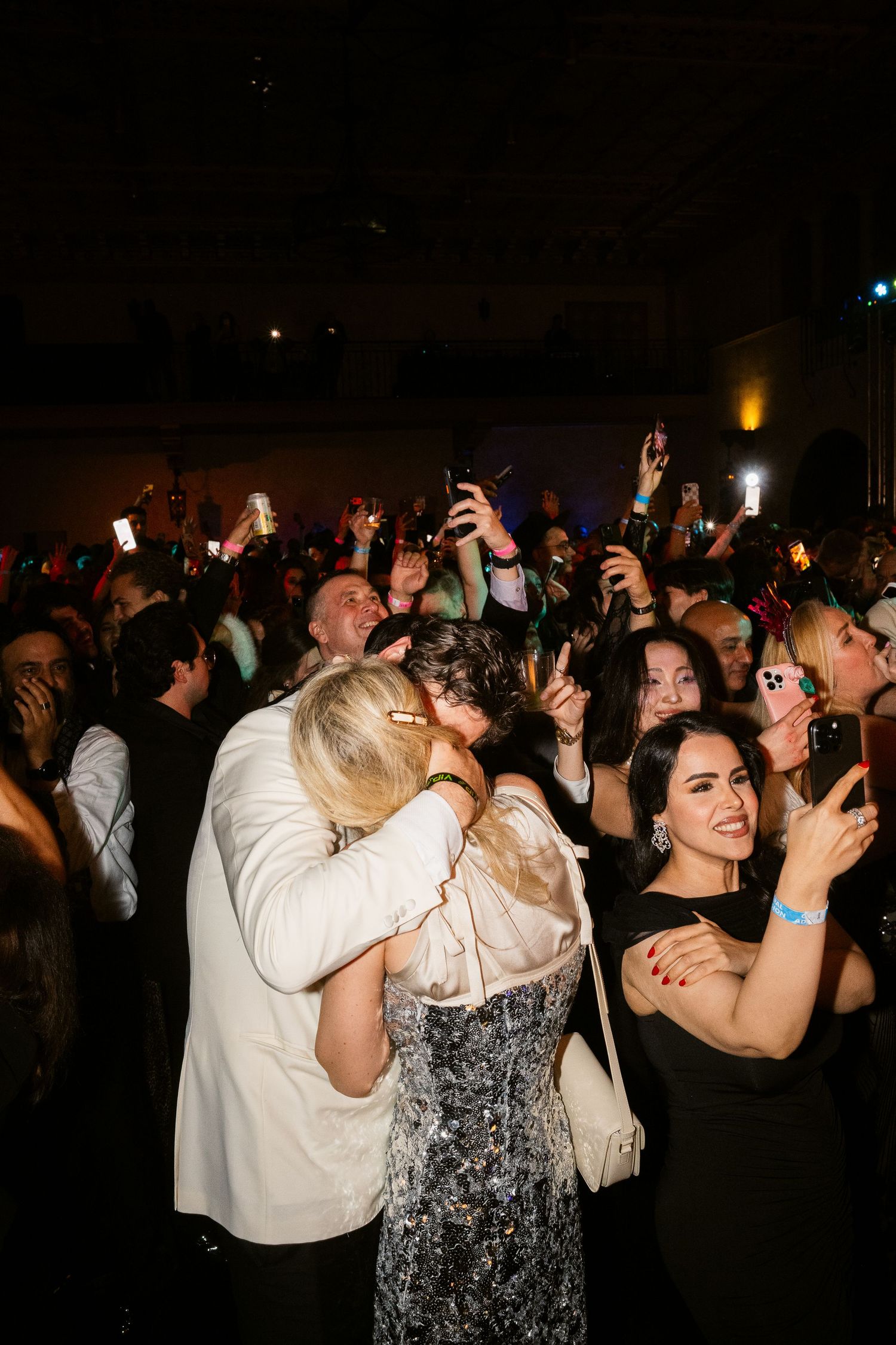 Couple sharing a kiss on a crowded dance floor at a formal event while guests celebrate and photograph the moment.