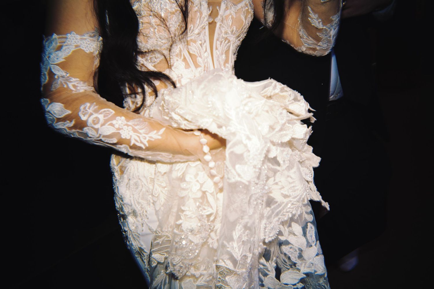 Close-up of a bride wearing an intricate white lace wedding gown with floral appliques and sheer sleeves.