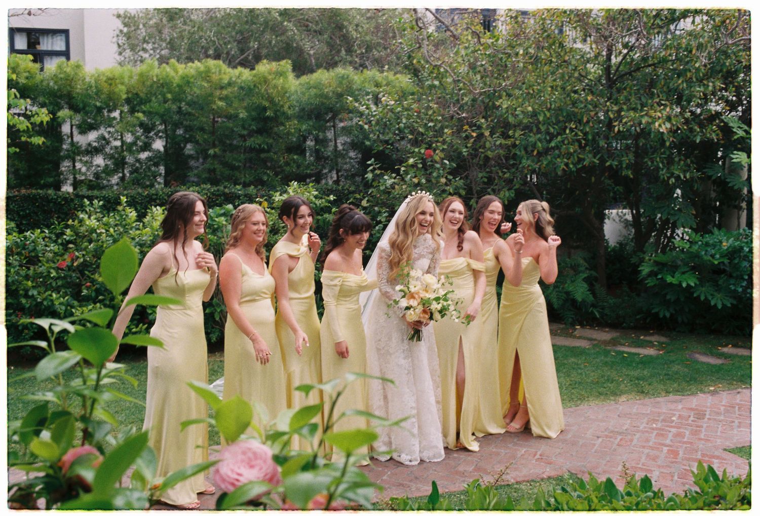 Bride in white gown with bridesmaids in yellow dresses walking through lush garden setting.
