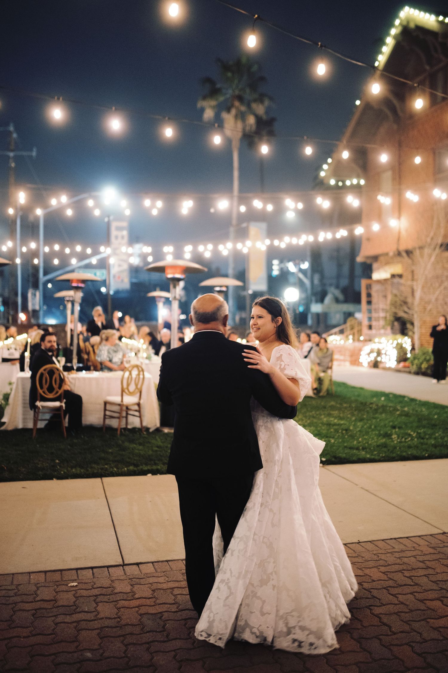 Bride and groom share first dance under glowing string lights at outdoor night wedding reception.