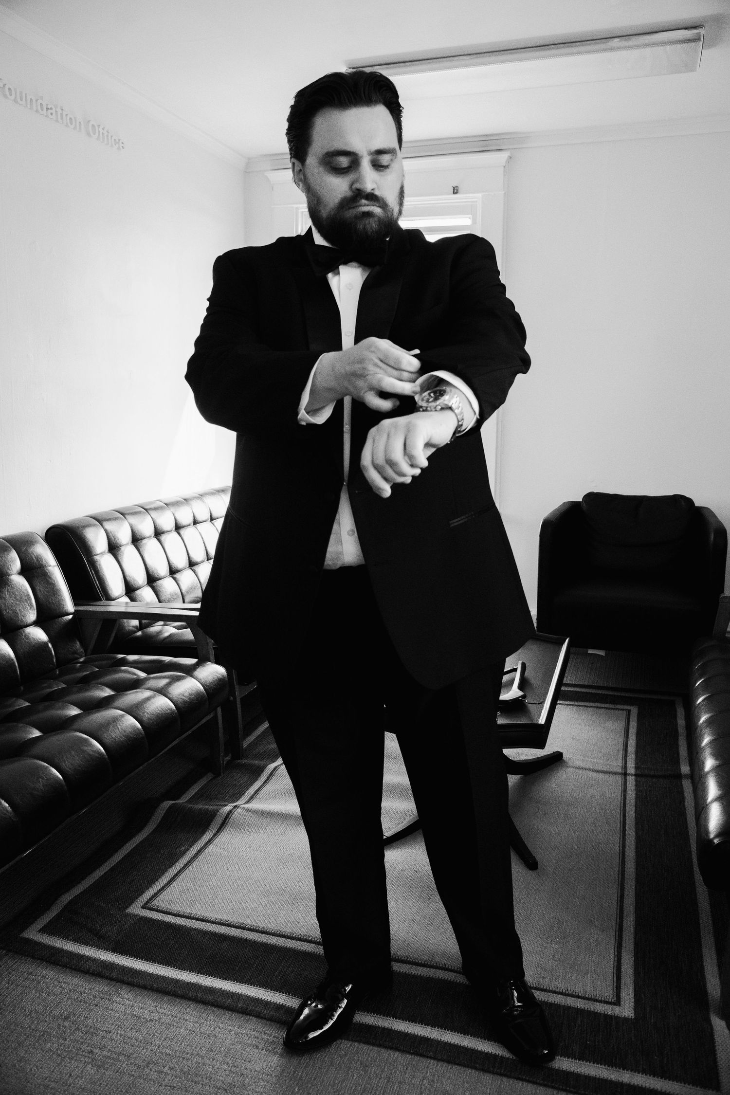 Groom in black tuxedo and bow tie checking watch while getting ready in room.