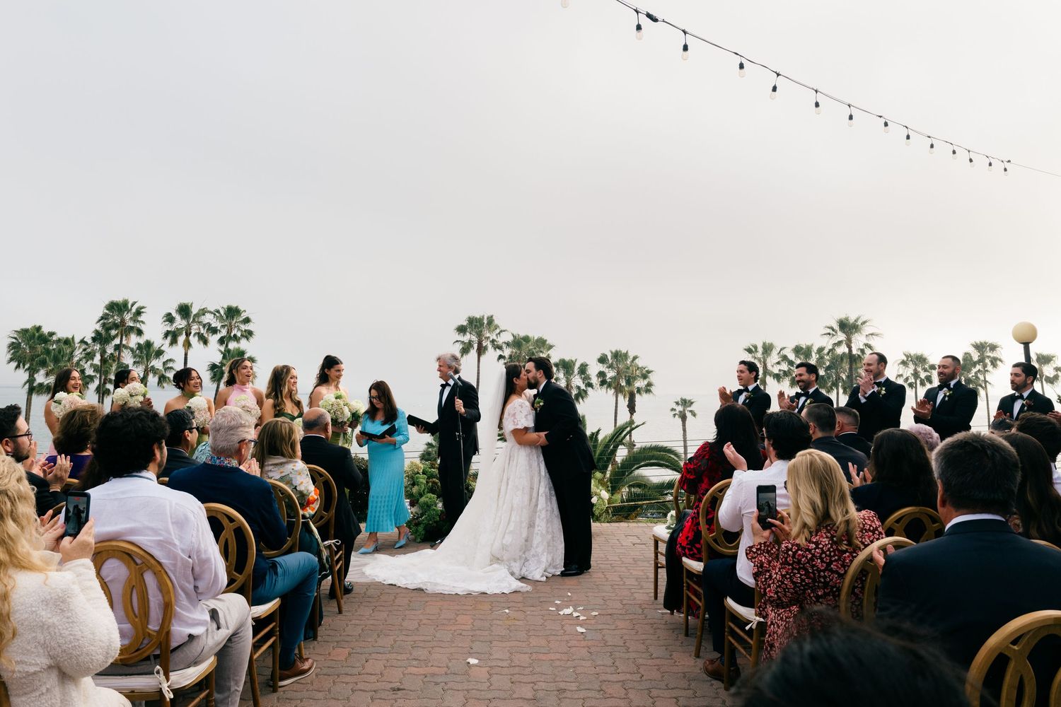 Bride and groom share first kiss at outdoor wedding ceremony with palm trees.