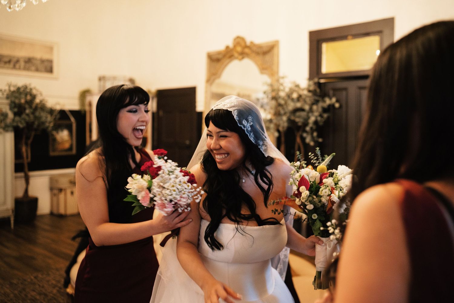 Excited bridesmaid reacts with joy beside smiling bride holding floral bouquets inside wedding venue.
