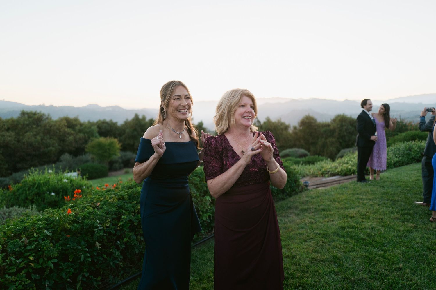 Two women smiling and laughing together at an outdoor wedding reception with mountain views at golden hour.