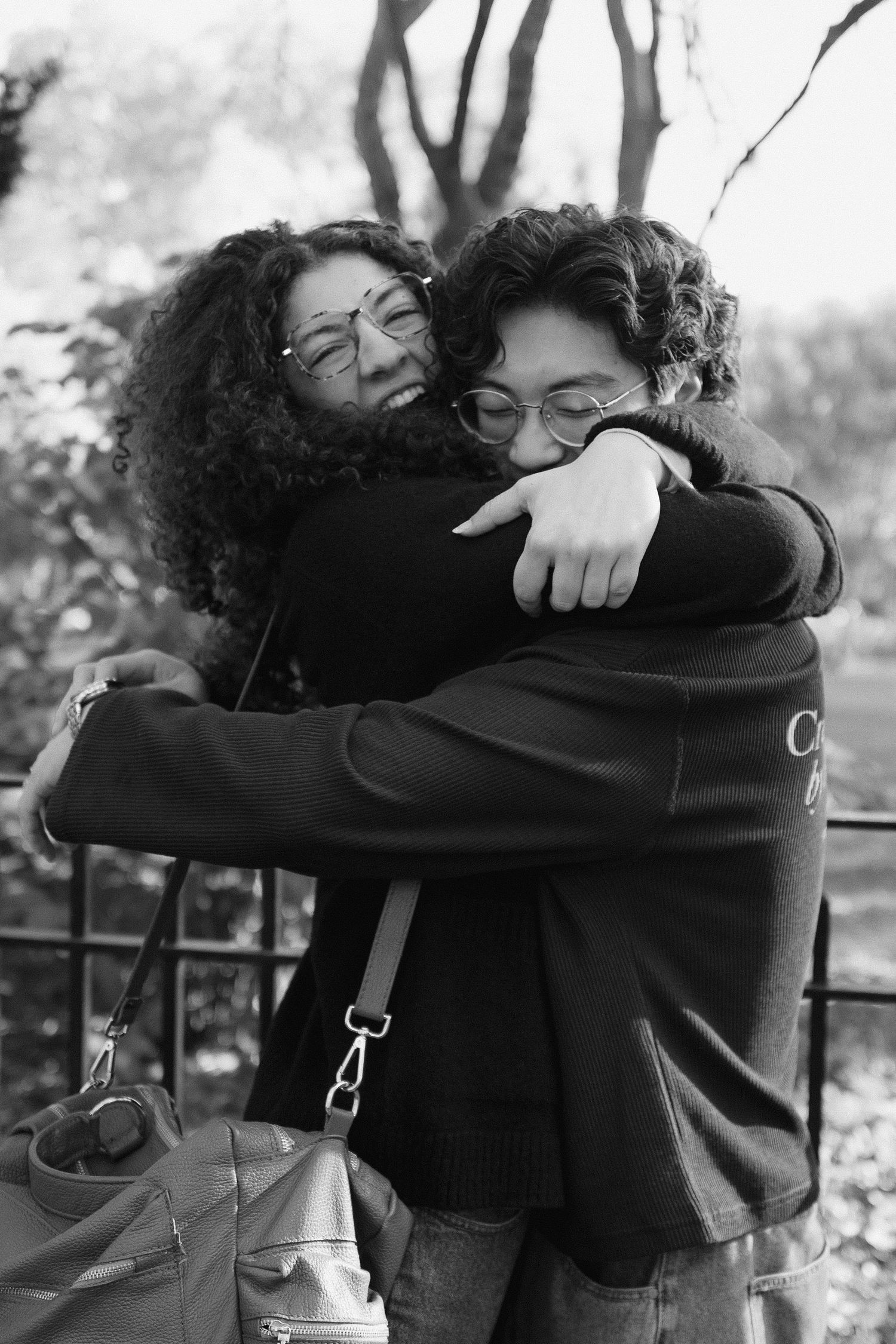 Two people sharing a warm embrace outdoors in a park, both wearing glasses and dark clothing.