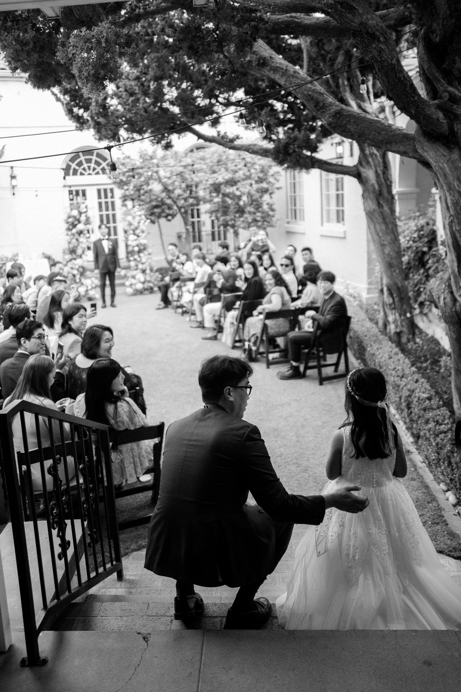 Black and white photo of bride and groom descending stairs toward outdoor wedding ceremony guests.