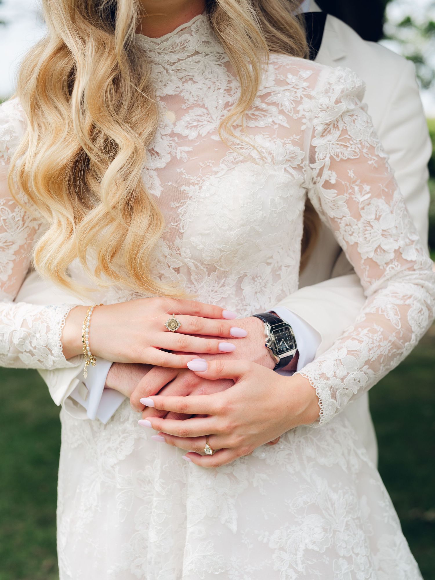 Close-up of bride and groom holding hands, showcasing lace wedding dress, gold bracelet, and luxury watch.